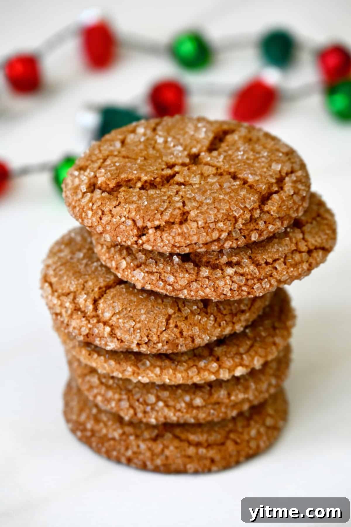 A delightful stack of sugar-dusted ginger cookies resting on a pristine white surface, complemented by festive red and green decorations in the background.