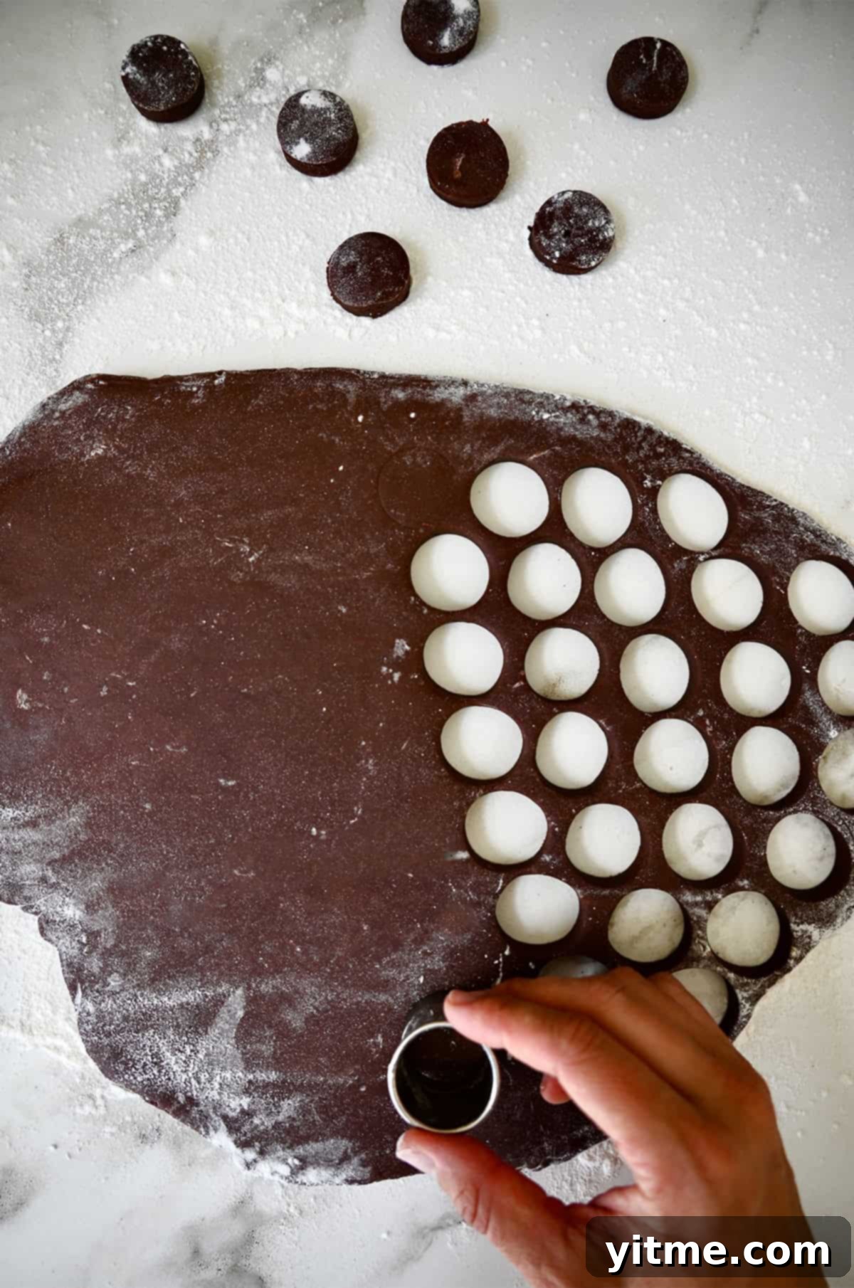 A baker's hands using a small round cookie cutter to cut out chocolate doughnut dough circles on a floured surface.
