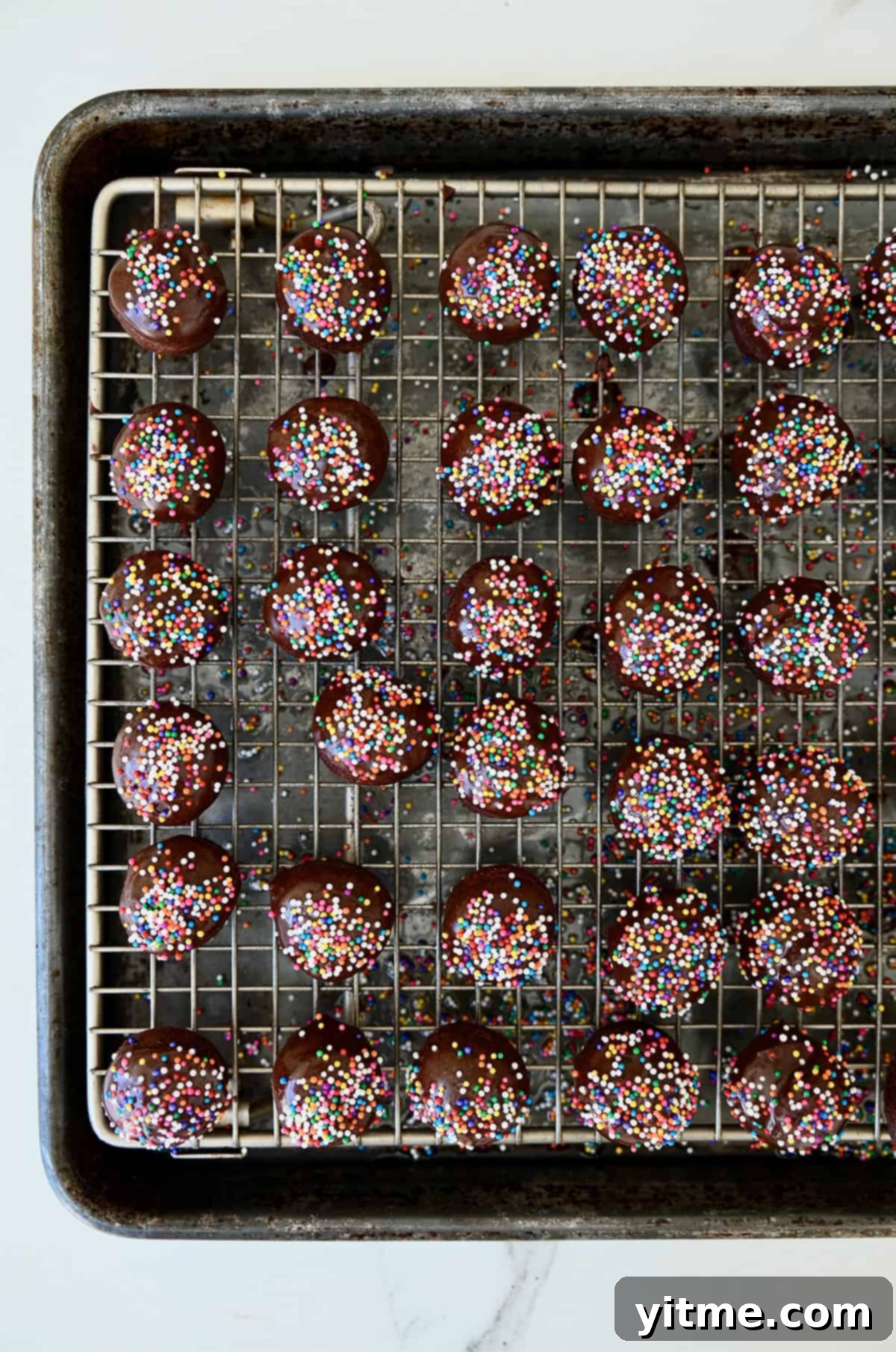Freshly glazed chocolate doughnut holes with colorful sprinkles drying on a wire rack.