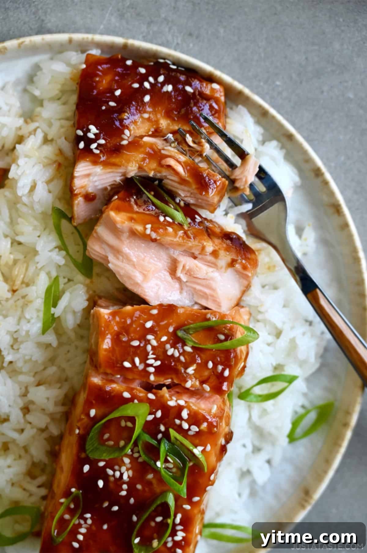 Baked honey garlic soy salmon garnished with sesame seeds and chopped scallions atop a bed of white rice on a plate with a fork.