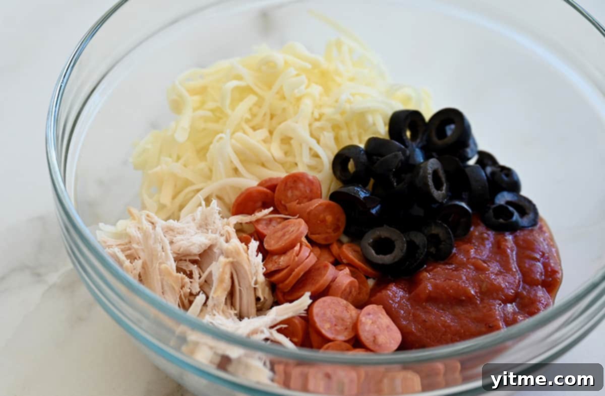 A clear bowl containing shredded chicken, mini pepperonis, black olives, shredded cheese and marinara sauce.