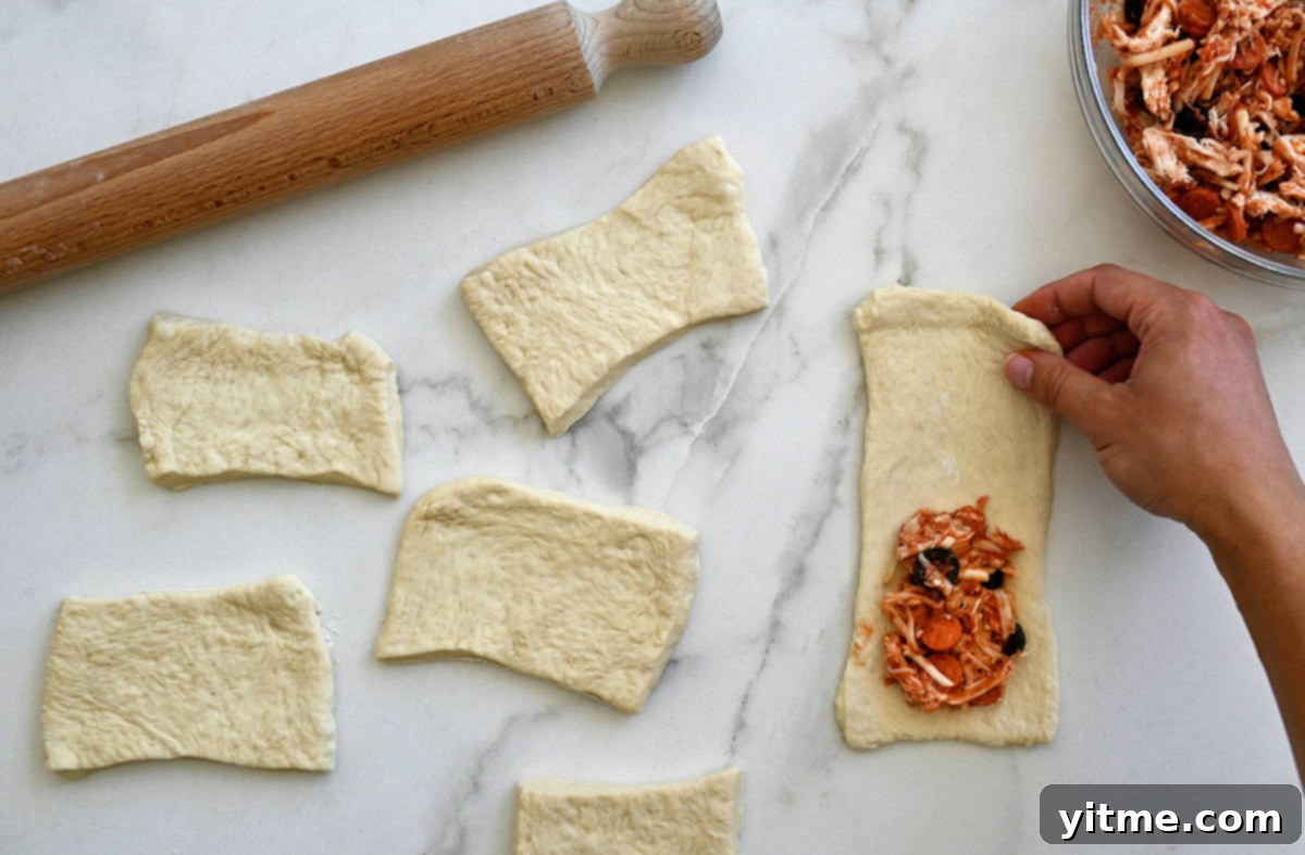 A rolling pin next to pieces of rectangular pizza dough with one piece topped with mini pepperoni, cheese and black olives.