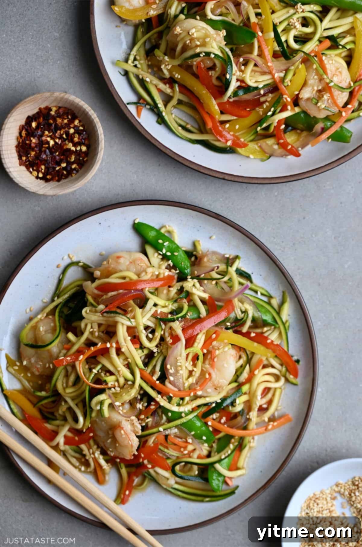 Zucchini noodle stir-fry with shrimp served on a plate next to crushed red pepper flakes.