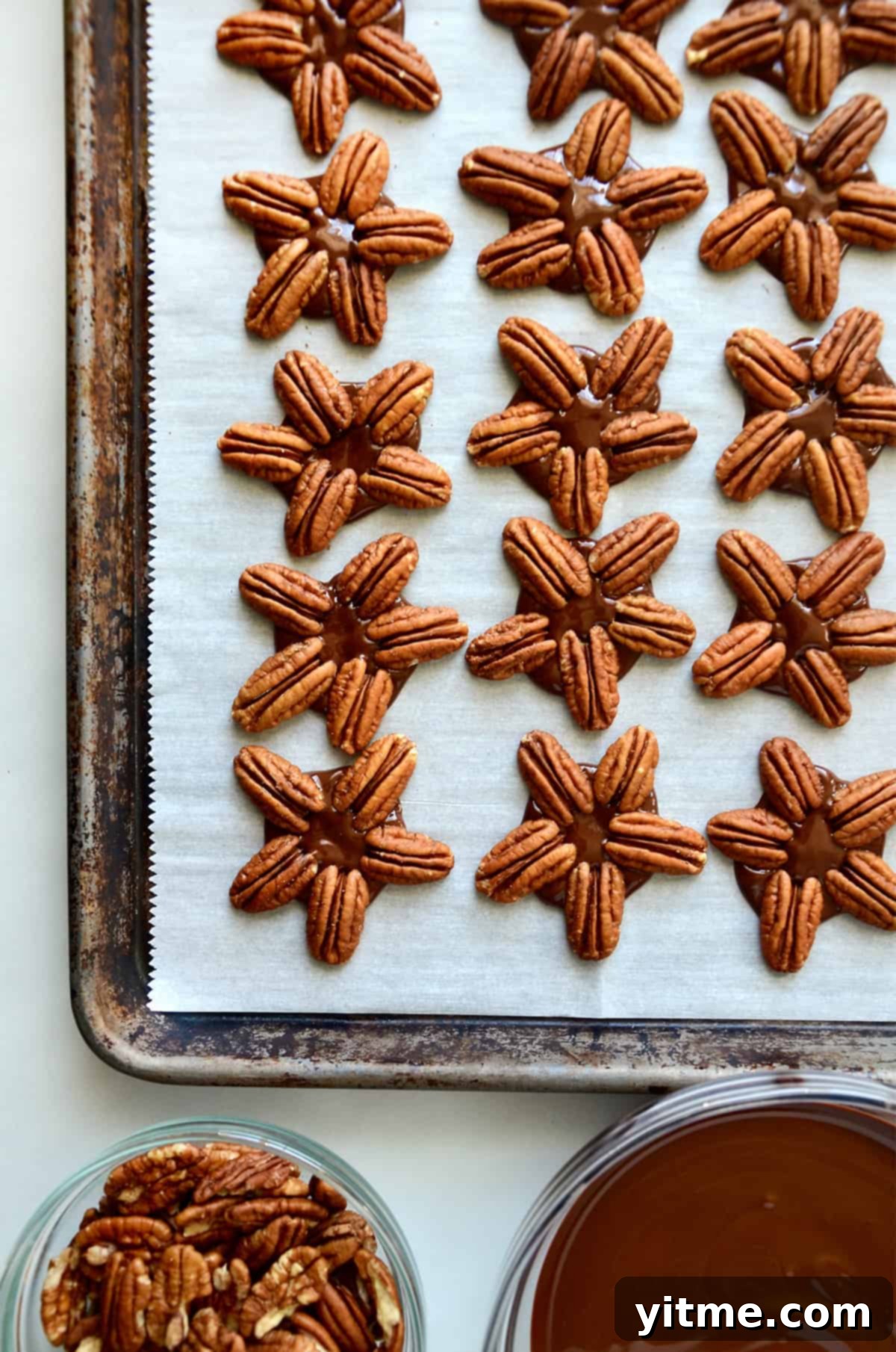 Chocolate turtles on a parchment-lined baking sheet with bowls of pecan halves and melted chocolate beside the baking sheet.