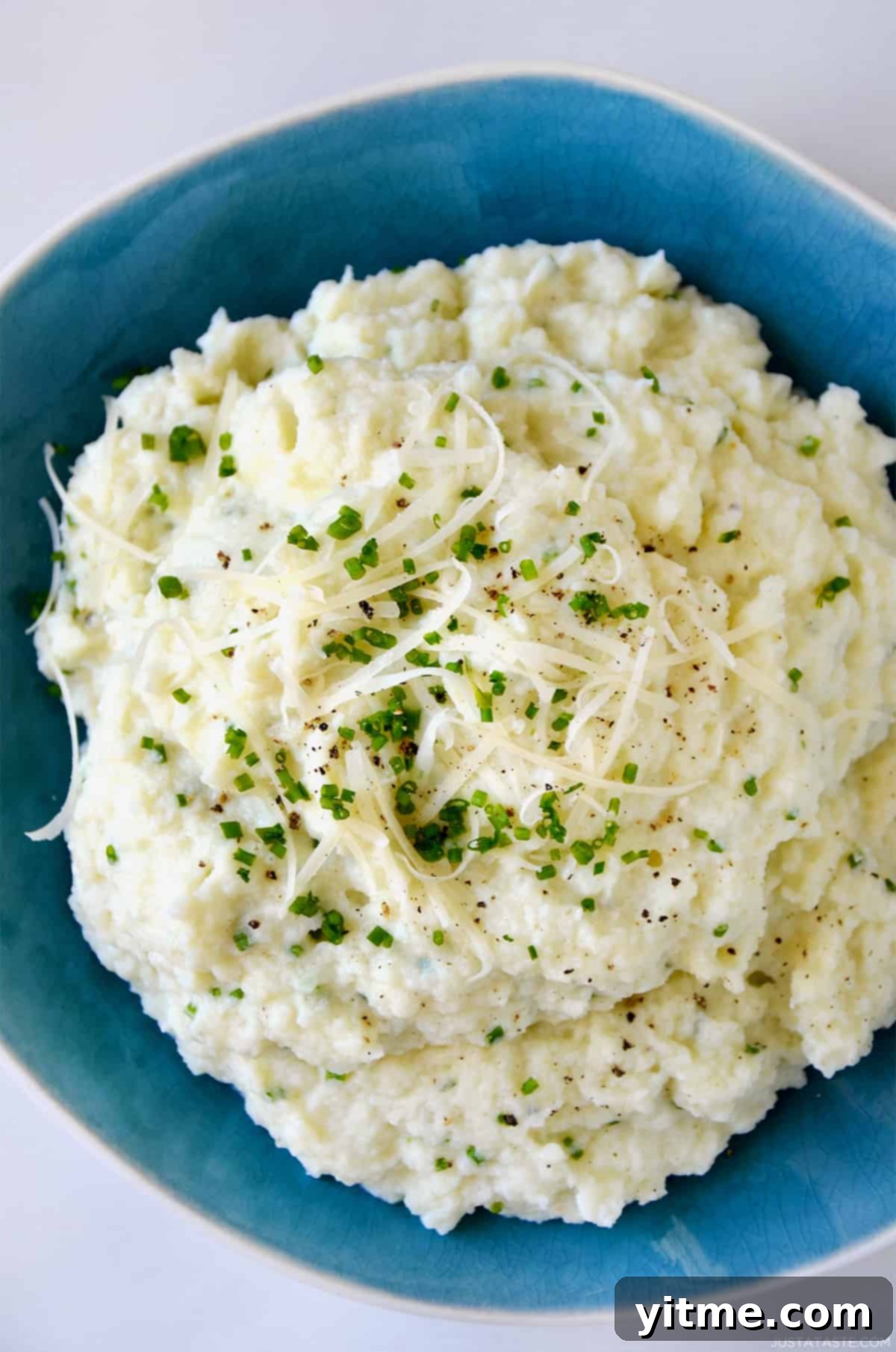 Cheesy Chive Mashed Cauliflower 2 Mashed cauliflower topped with shredded cheese, finely minced chives and black pepper in a blue bowl.