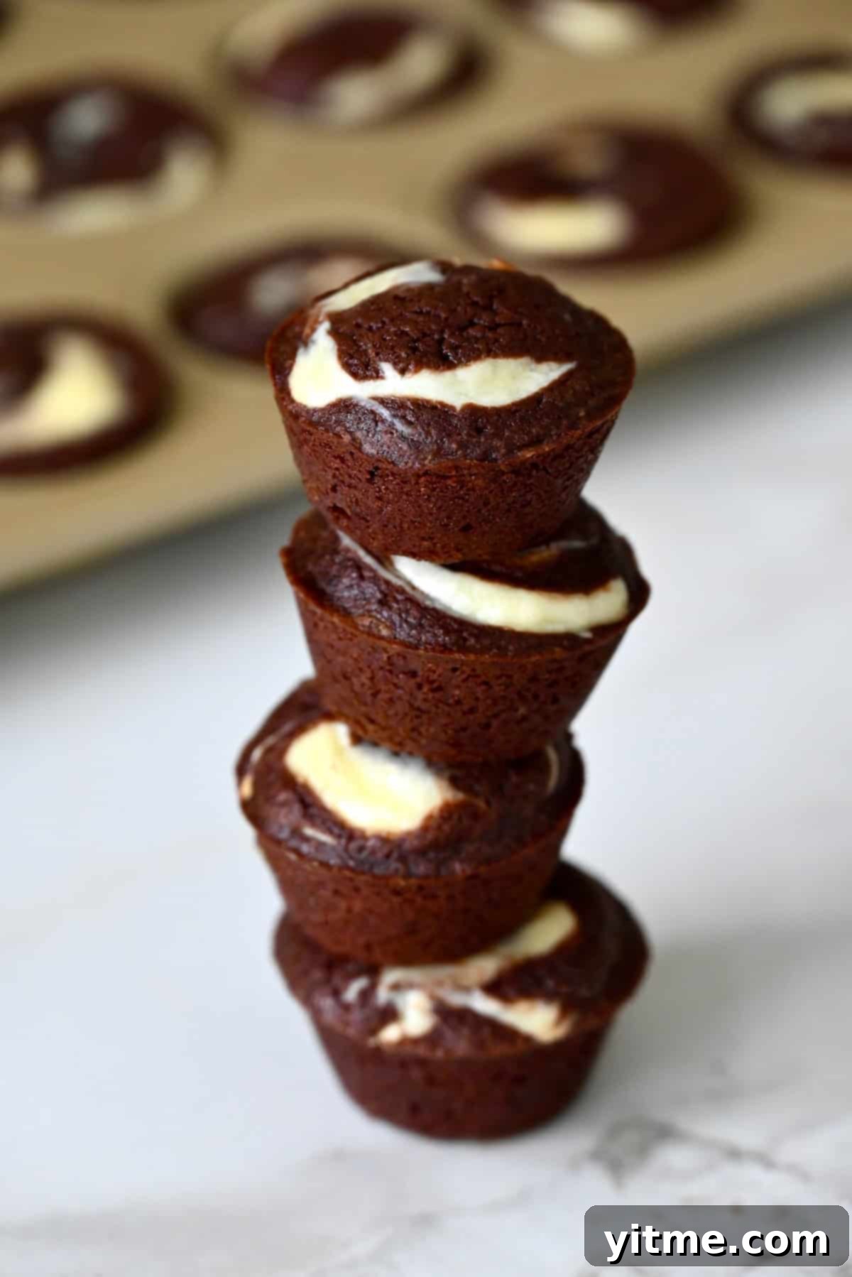 Cheesecake brownie bites are stacked on a marble counter, with a mini muffin pan in the background.