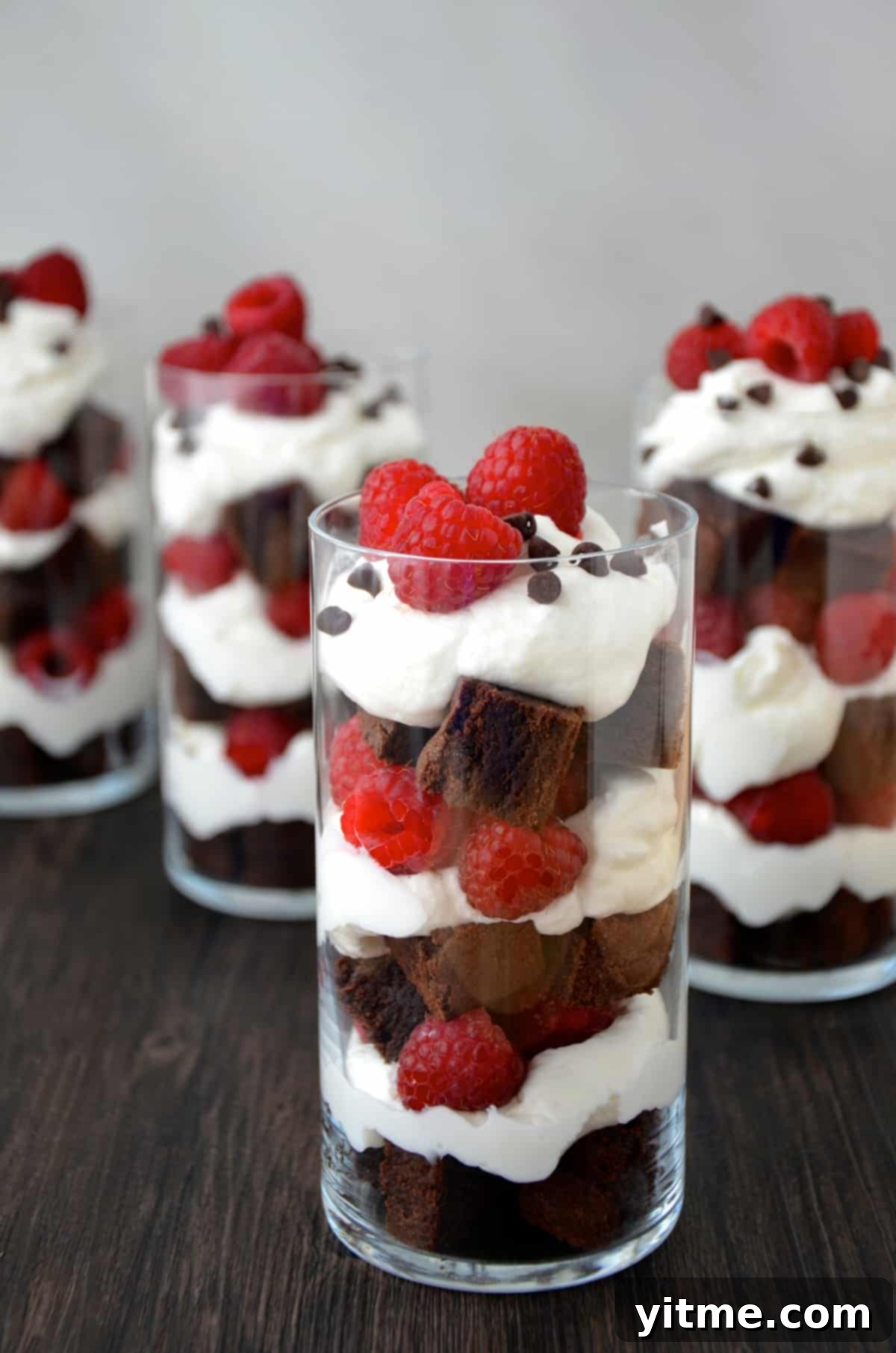 Several parfait glasses filled with layers of brownie pieces, whipped cream and fresh raspberries.