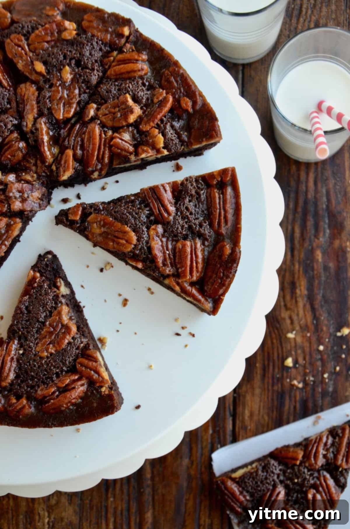 Slices of Pecan Pie Brownies sit on a white scalloped cake stand. Nearby is a glass of milk with two striped straws.