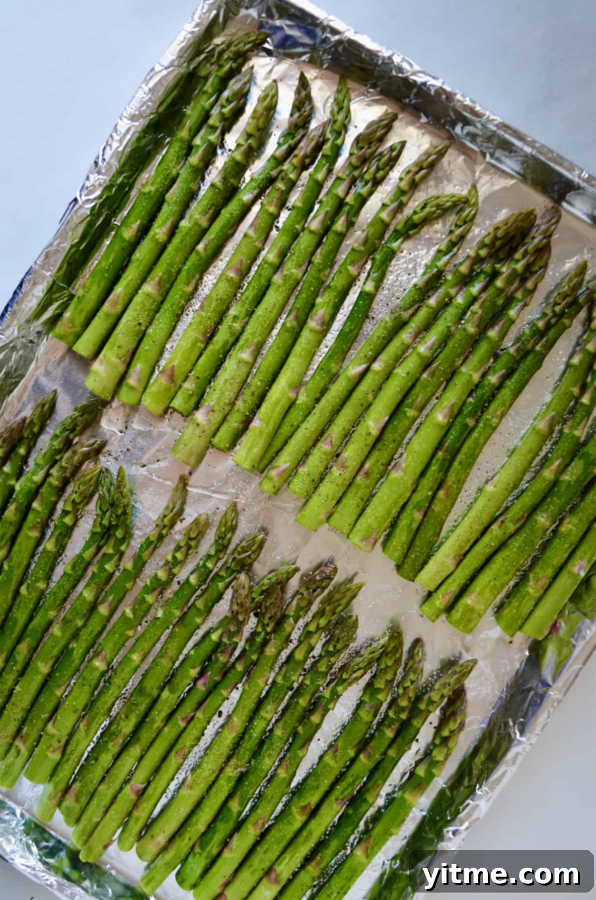 Two rows of asparagus seasoned with salt and pepper on a foil-lined baking sheet, ready for roasting.