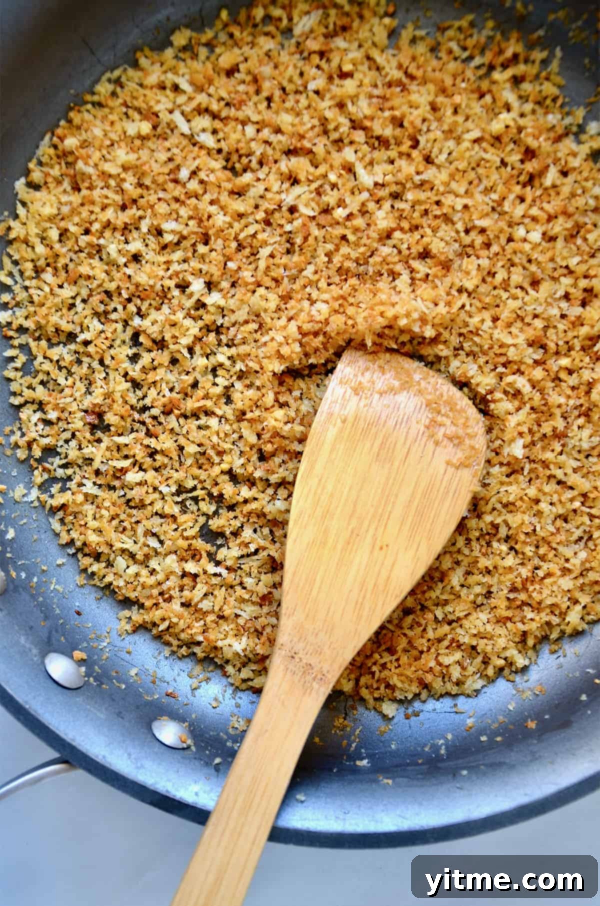 Toasted Panko breadcrumbs, golden and crispy, shown in a nonstick skillet with a spoon resting against its side.