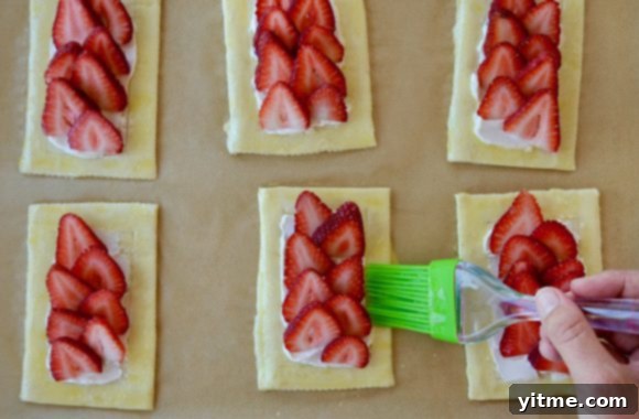 Hand with brush brushing egg wash on pastry with strawberries 