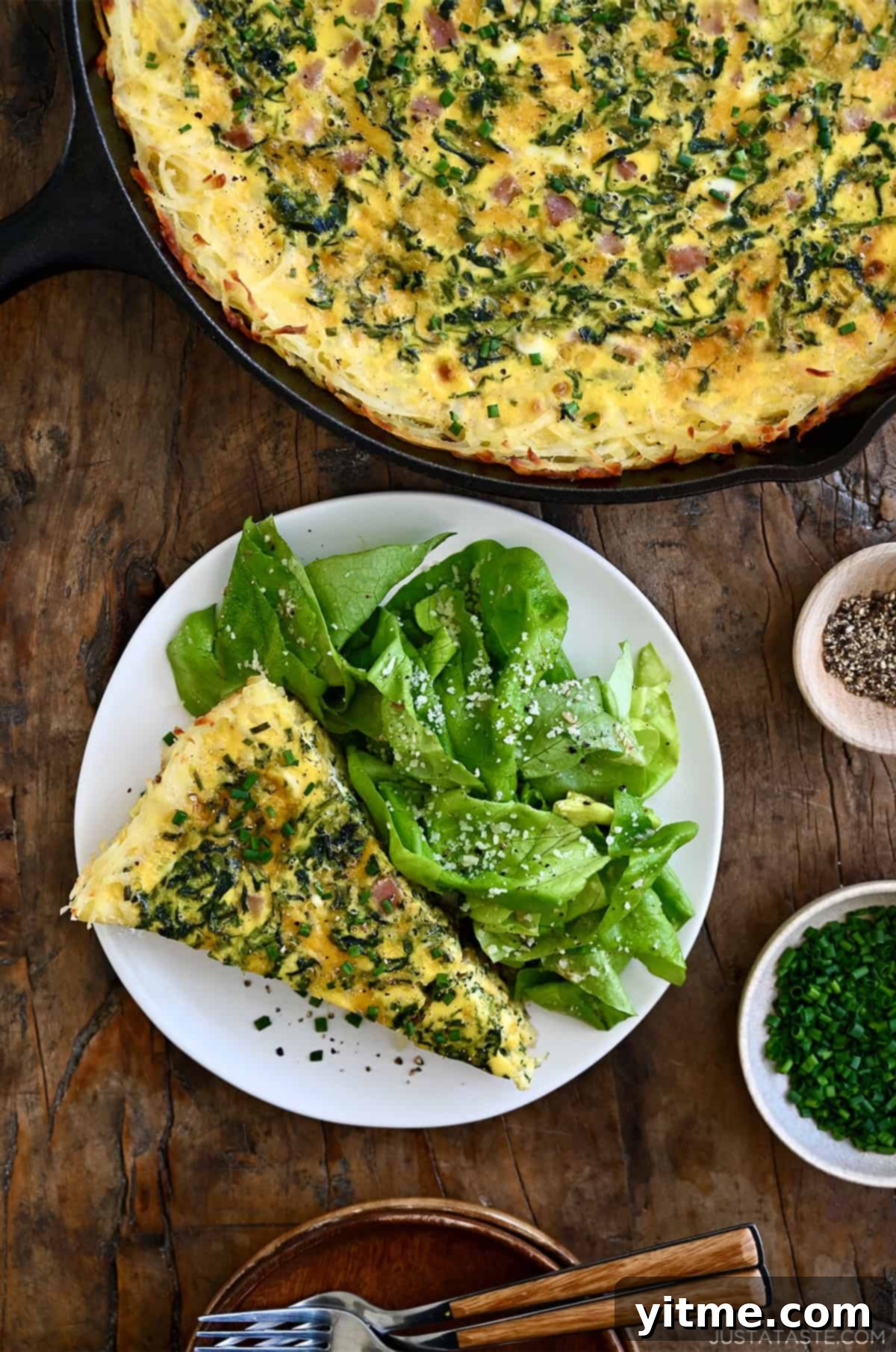 A slice of ham and cheese quiche with a crispy hash brown crust served on a plate with dressed greens.