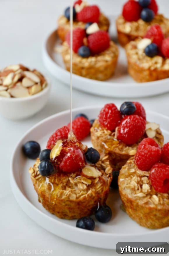 Baked Oatmeal Cups on a white plate topped with fresh raspberries and blueberries being drizzled with warm maple syrup.