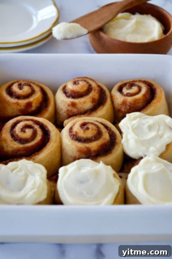 A white baking dish filled with freshly baked cinnamon rolls, accompanied by a bowl of creamy frosting.