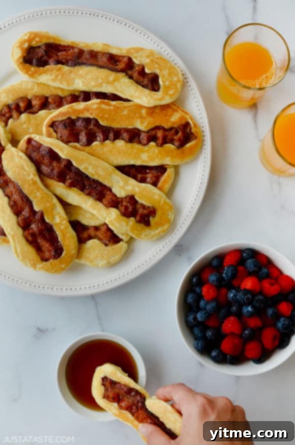 White plate containing Bacon Pancake Dippers next to glasses of orange juice, small bowl with berries and hand dunking pancake dipper in maple syrup.