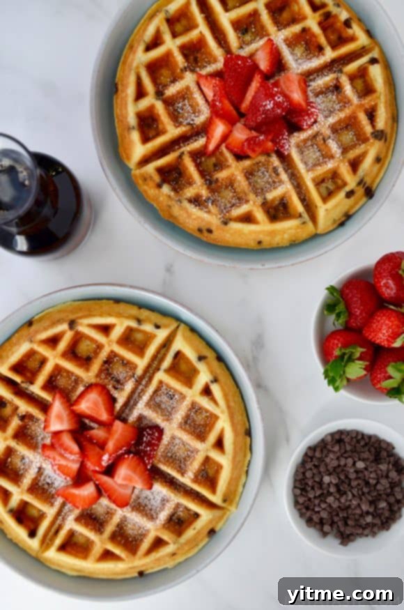 Buttermilk Chocolate Chip Waffles topped with strawberries and powdered sugar on pale blue plates next to a small white bowl filled with chocolate chips, a small glass pitcher with maple syrup and a small white bowl with strawberries.