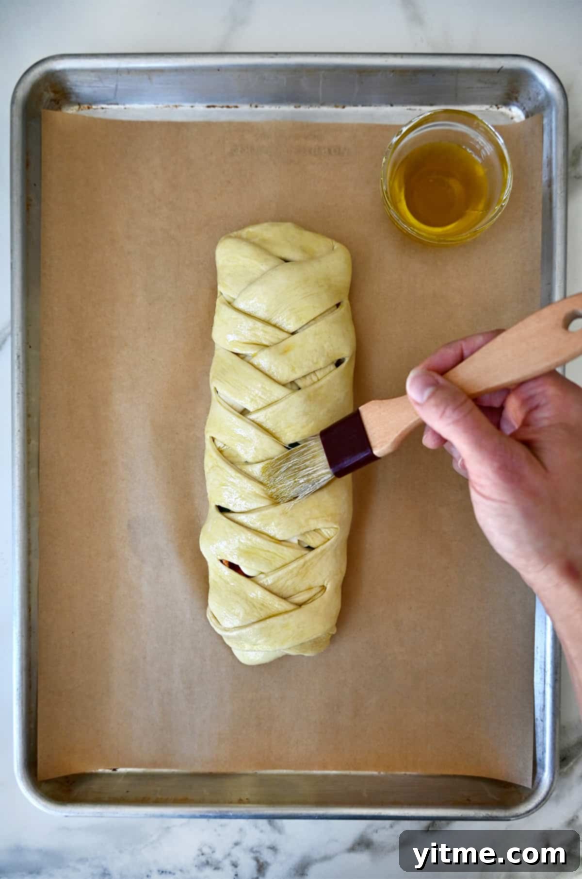 A hand holding a pastry brush applies olive oil to a braided pizza bread on a parchment paper-lined baking sheet.