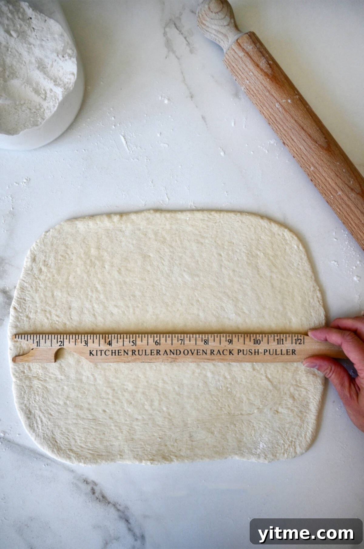A hand holding a ruler over pizza dough rolled into a 13'' rectangle. 