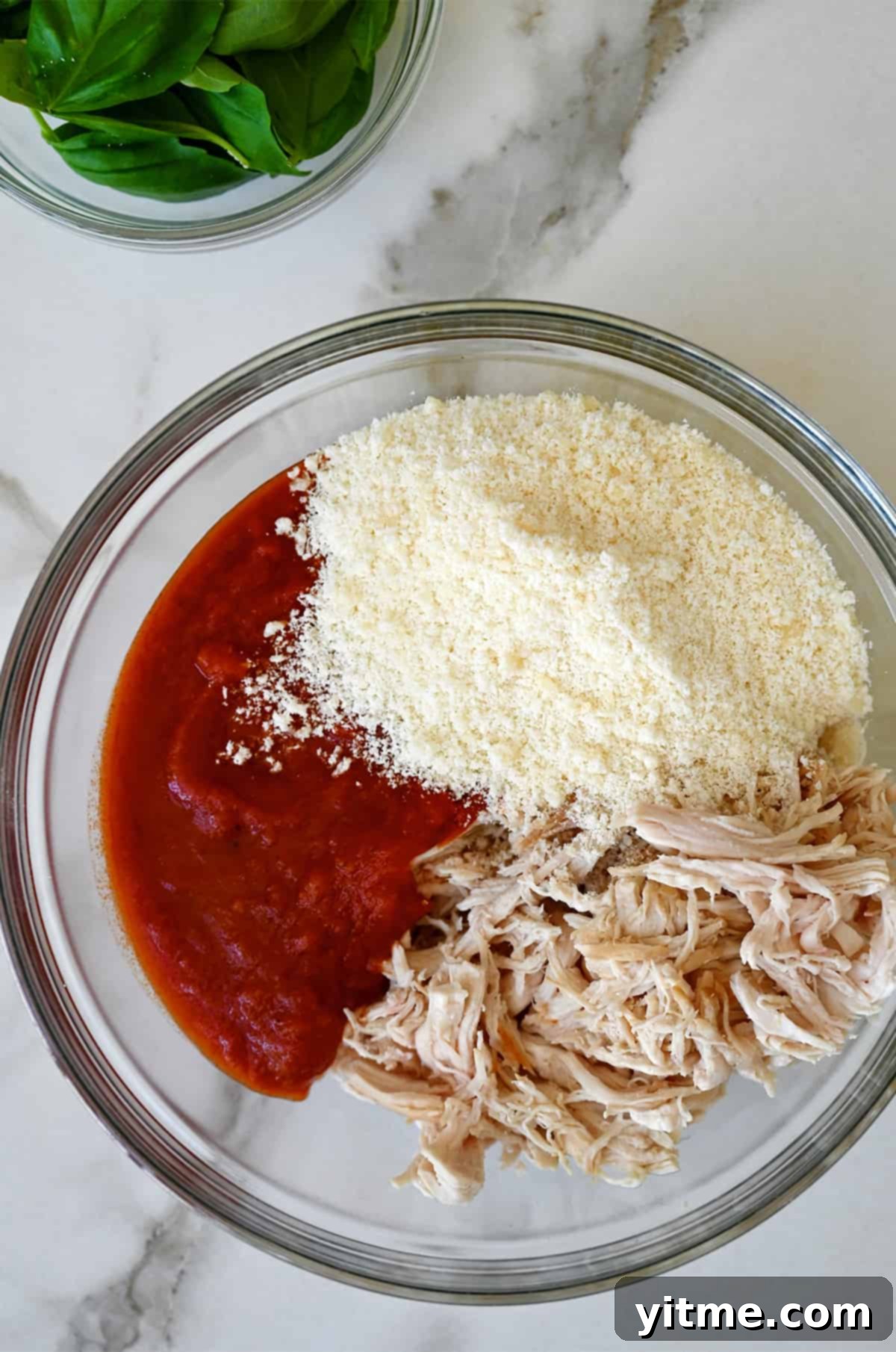 A glass bowl containing shredded chicken, marinara sauce and grated Parmesan cheese.