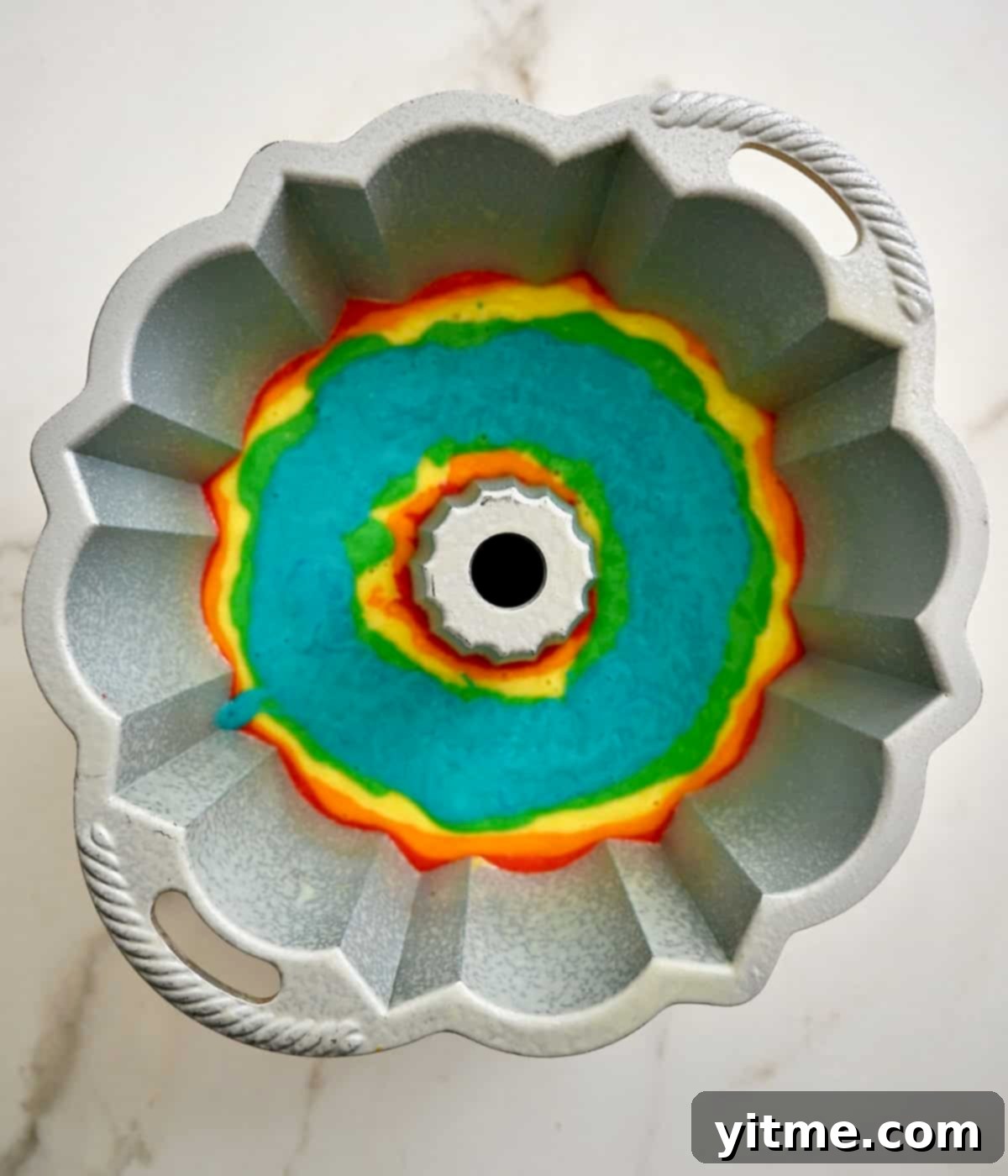 Blue cake layer atop green, yellow, orange and red cake layers in a bundt pan, demonstrating the ongoing delicate layering.