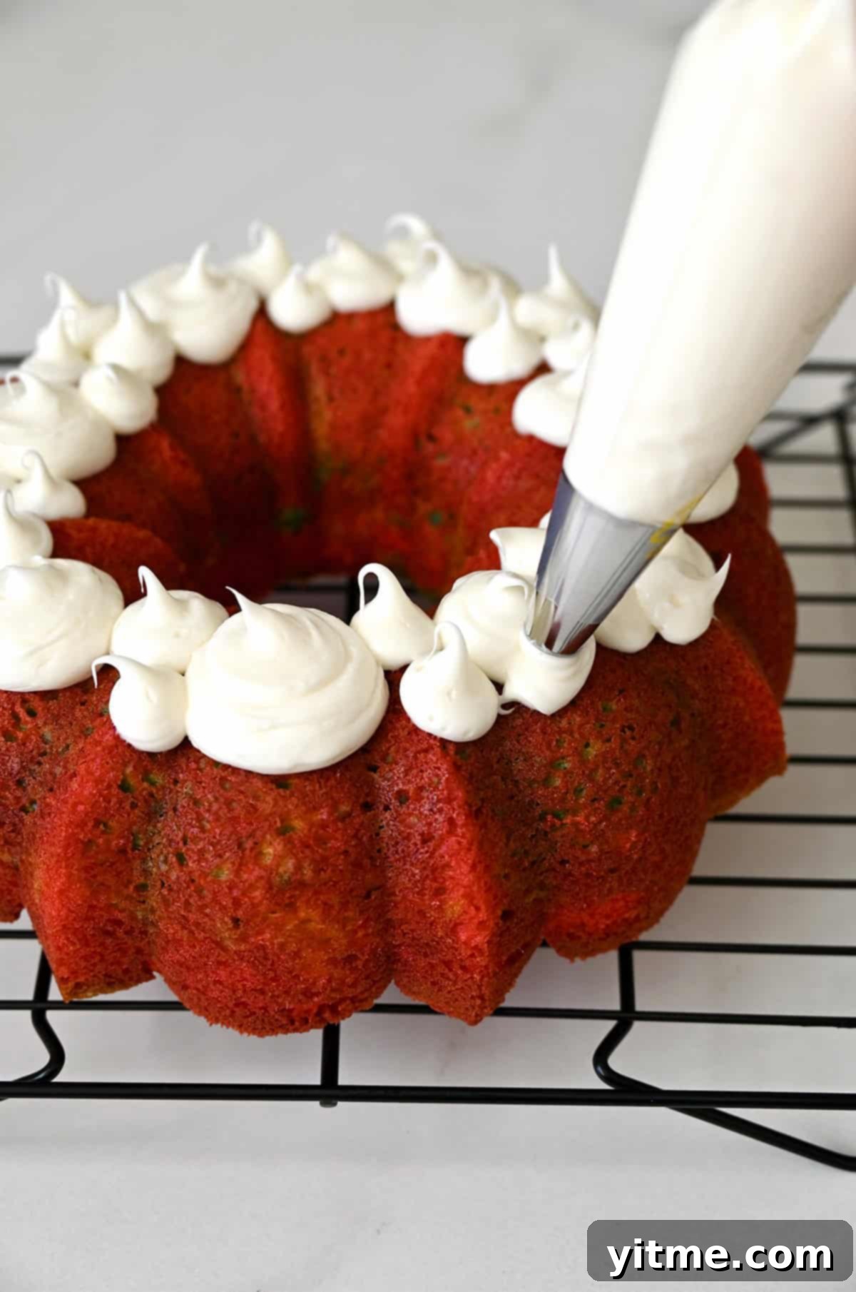 Piping vanilla frosting onto a rainbow cake on a wire cooling rack, showing the decorative process.