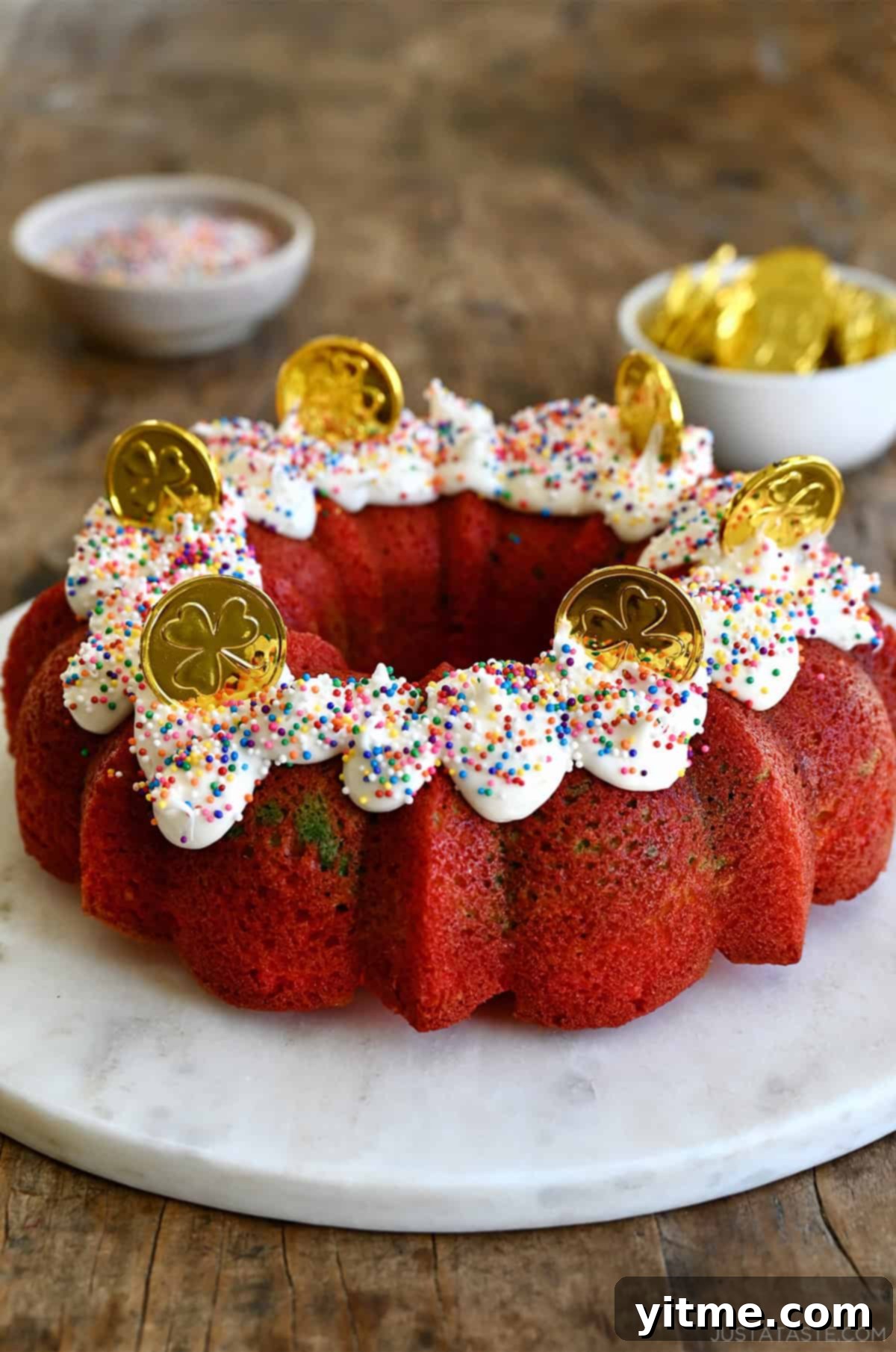 Rainbow cake topped with vanilla frosting, rainbow sprinkles and milk chocolate coins on a round cake platter, ready to be served.