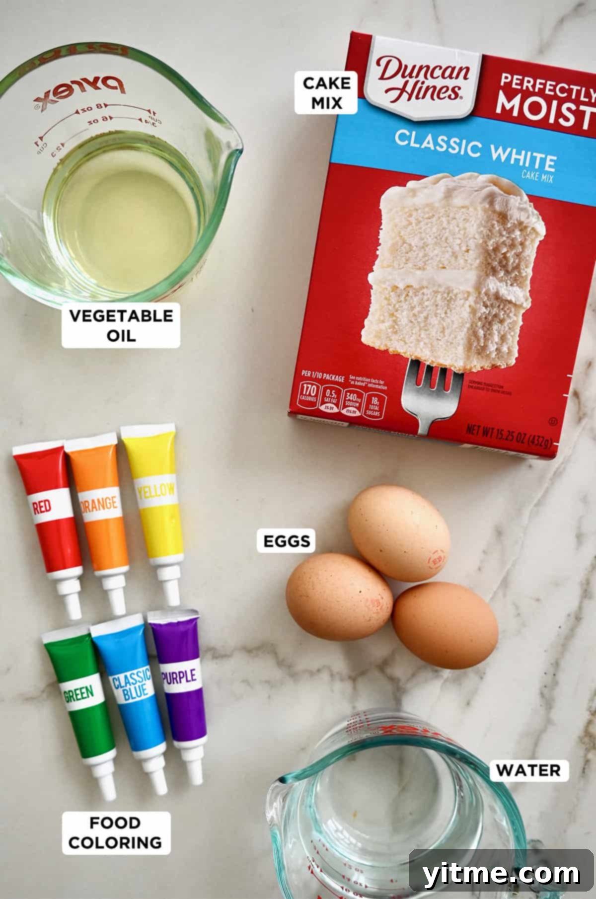 Rainbow cake ingredients: A box of white cake mix, vegetable oil, eggs, food coloring and water, neatly arranged.