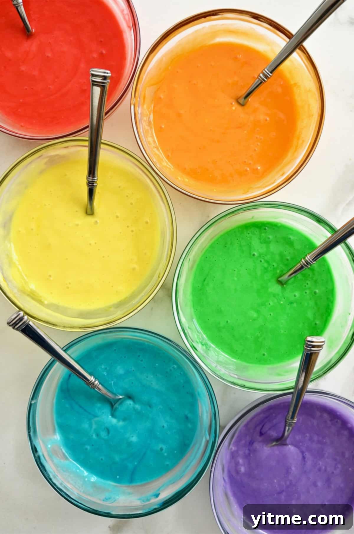 Six bowls containing cake batter dyed the colors of the rainbow: red, orange, yellow, green, blue and purple, ready for layering.