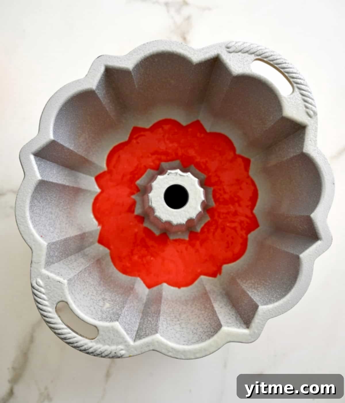 A bundt pan containing the first layer of red cake batter, forming the base of the rainbow cake.