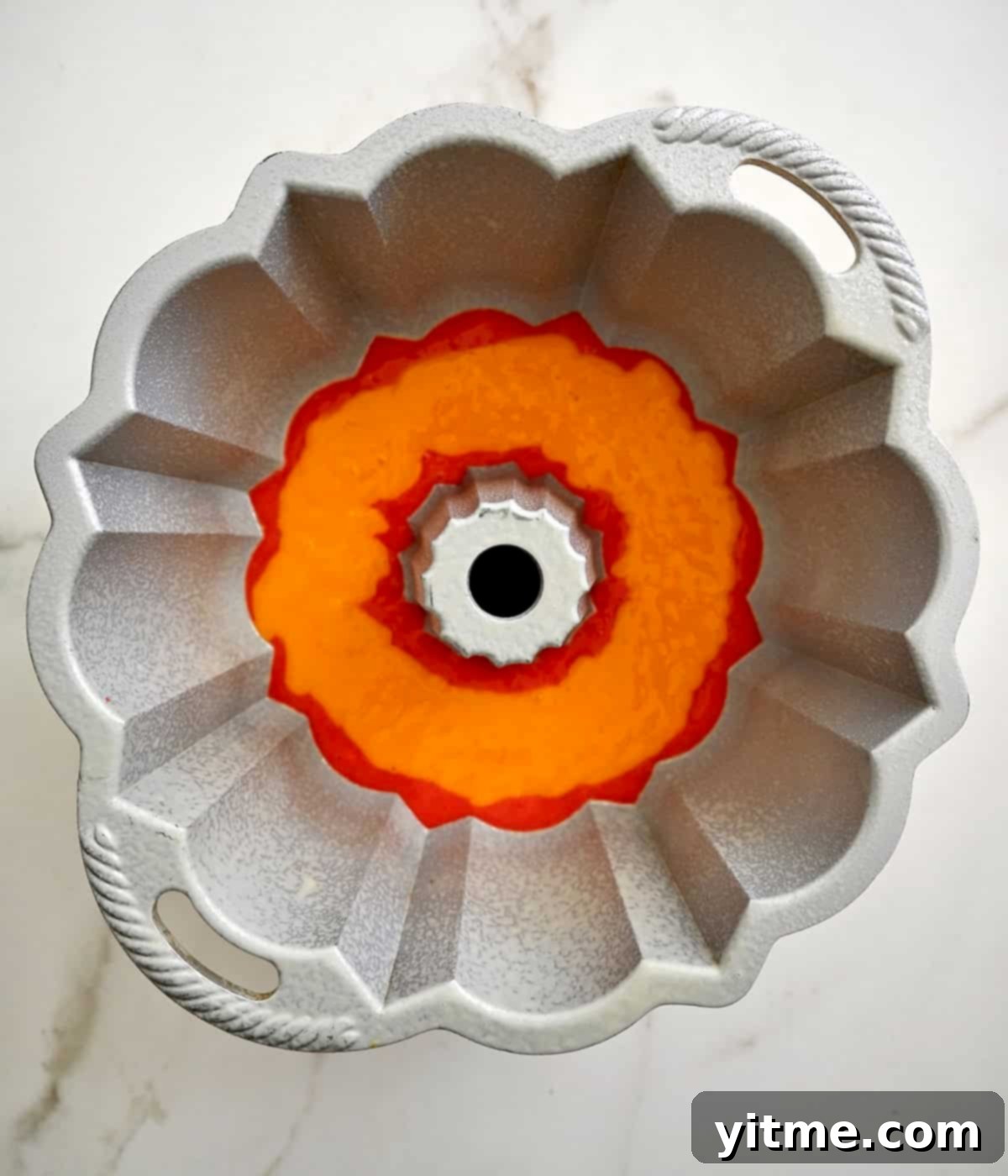 Orange layer of a rainbow bundt cake carefully poured atop the red cake batter, maintaining distinct separation.