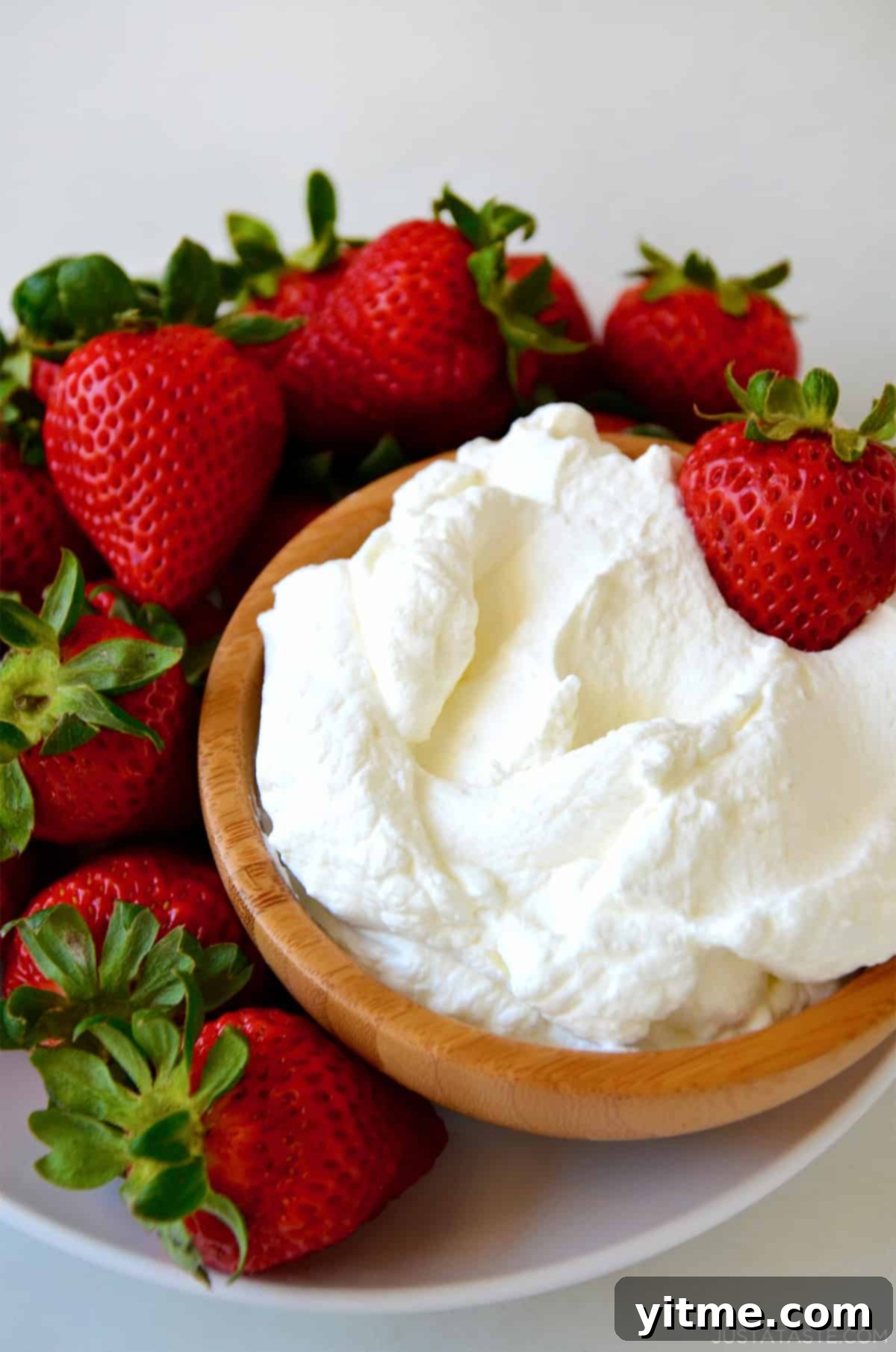 Homemade whipped cream in a bowl on a plate surrounded by strawberries, showcasing its fluffy and stable texture.