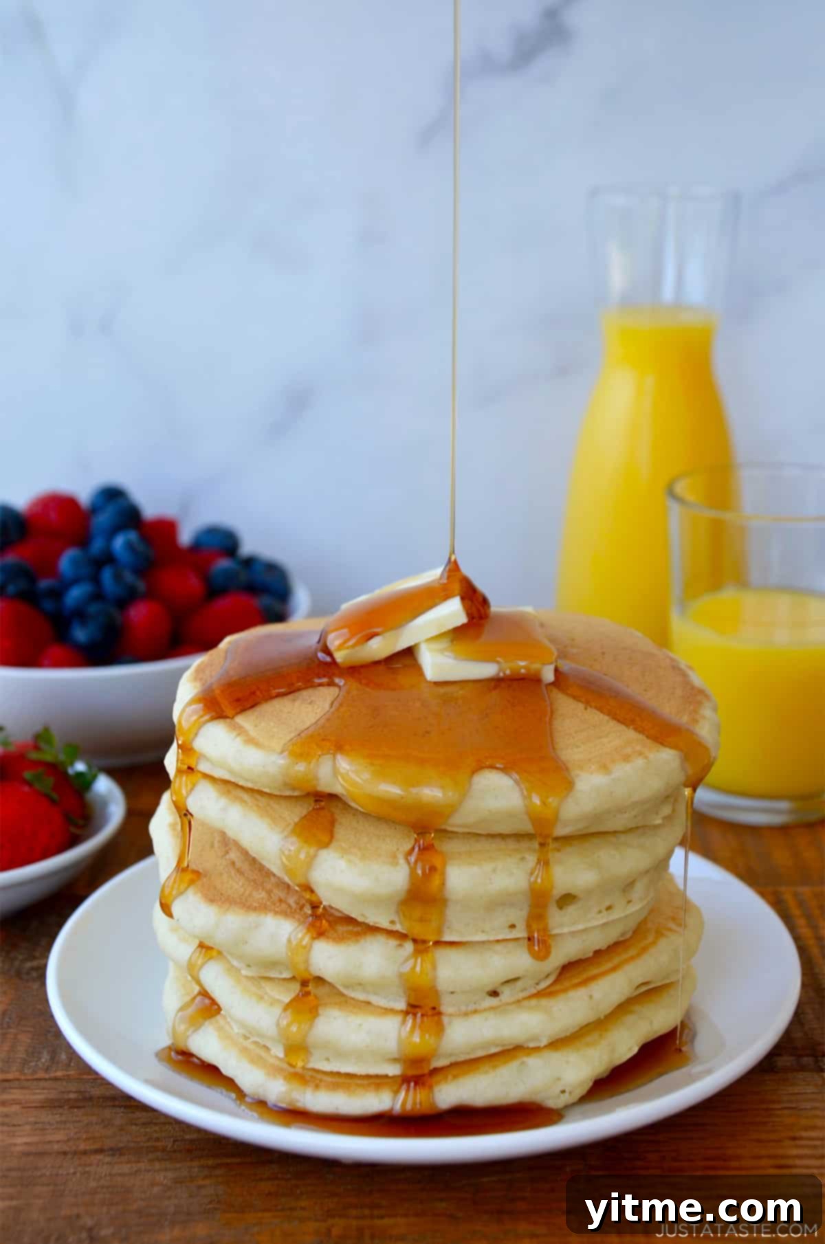 A tall stack of buttermilk pancakes dripping with maple syrup and topped with two pads of butter. Orange juice and fruit are in the background.