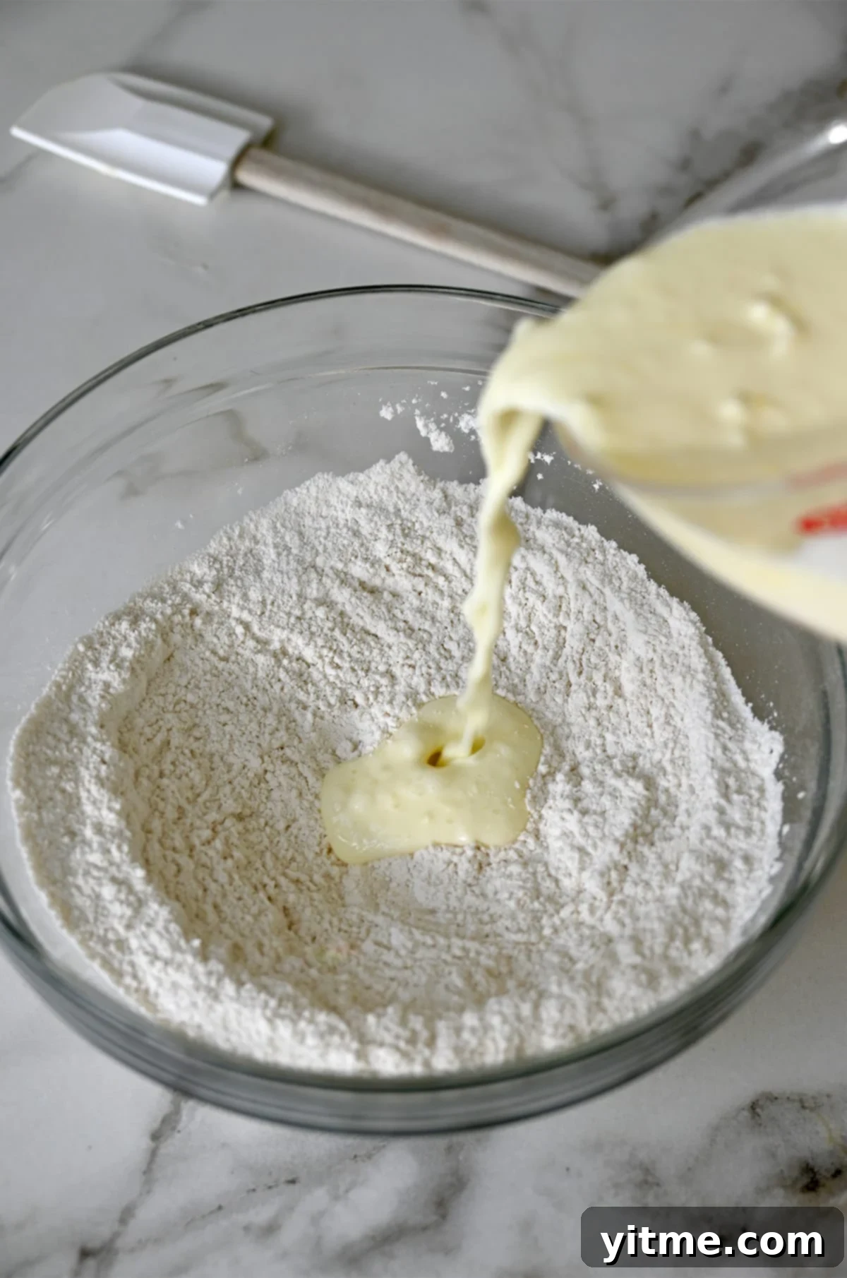 Buttermilk from a liquid measuring cup being poured atop flour in a glass bowl.