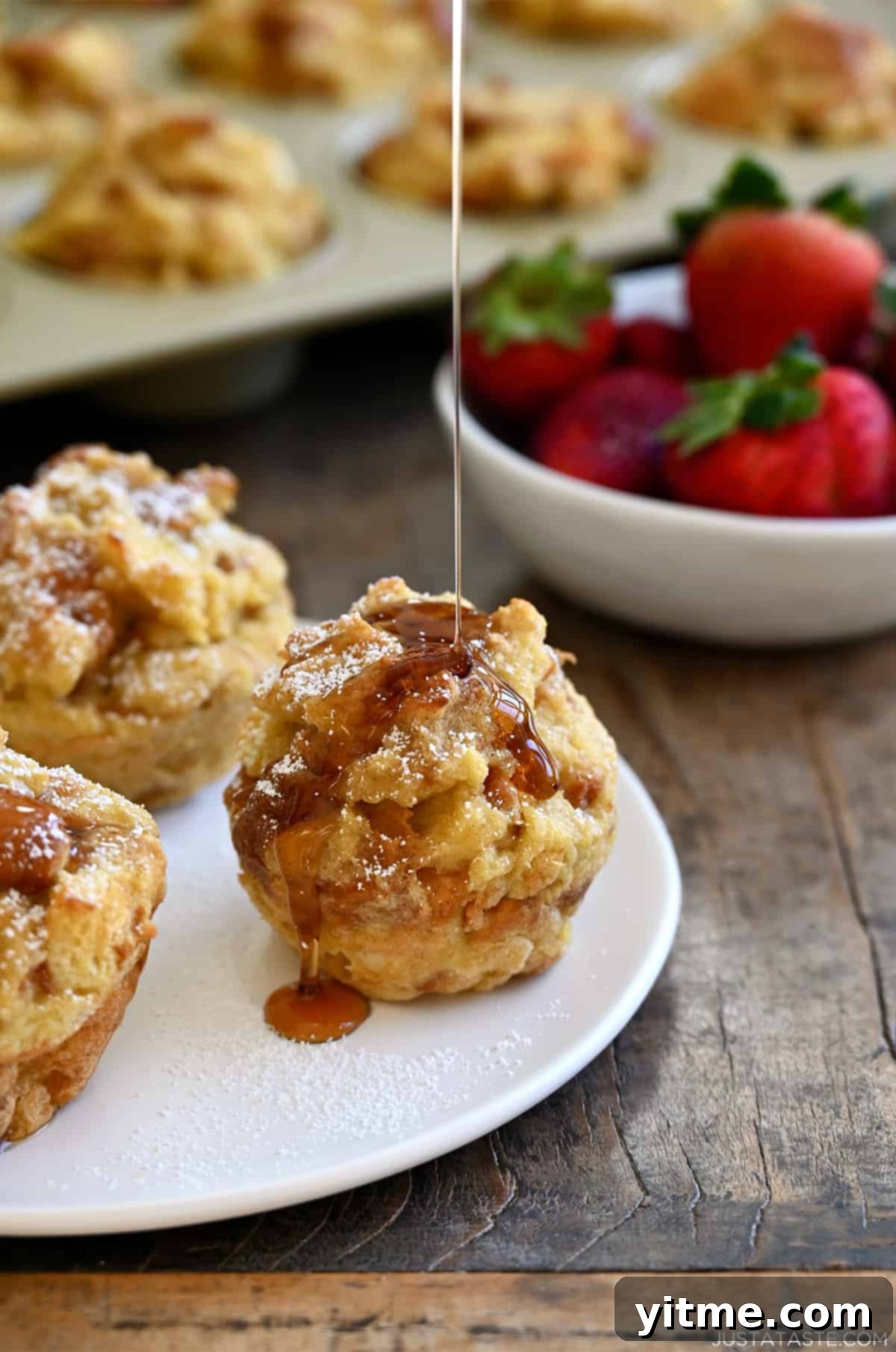 Cinnamon-Swirl French Toast Bites 6 A French toast muffin on a white plate being drizzled with maple syrup with a bowl of strawberries.