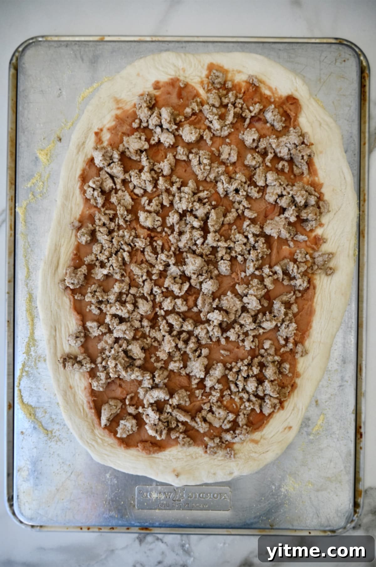 Pizza dough topped with refried beans and cooked ground chicken on a baking sheet.