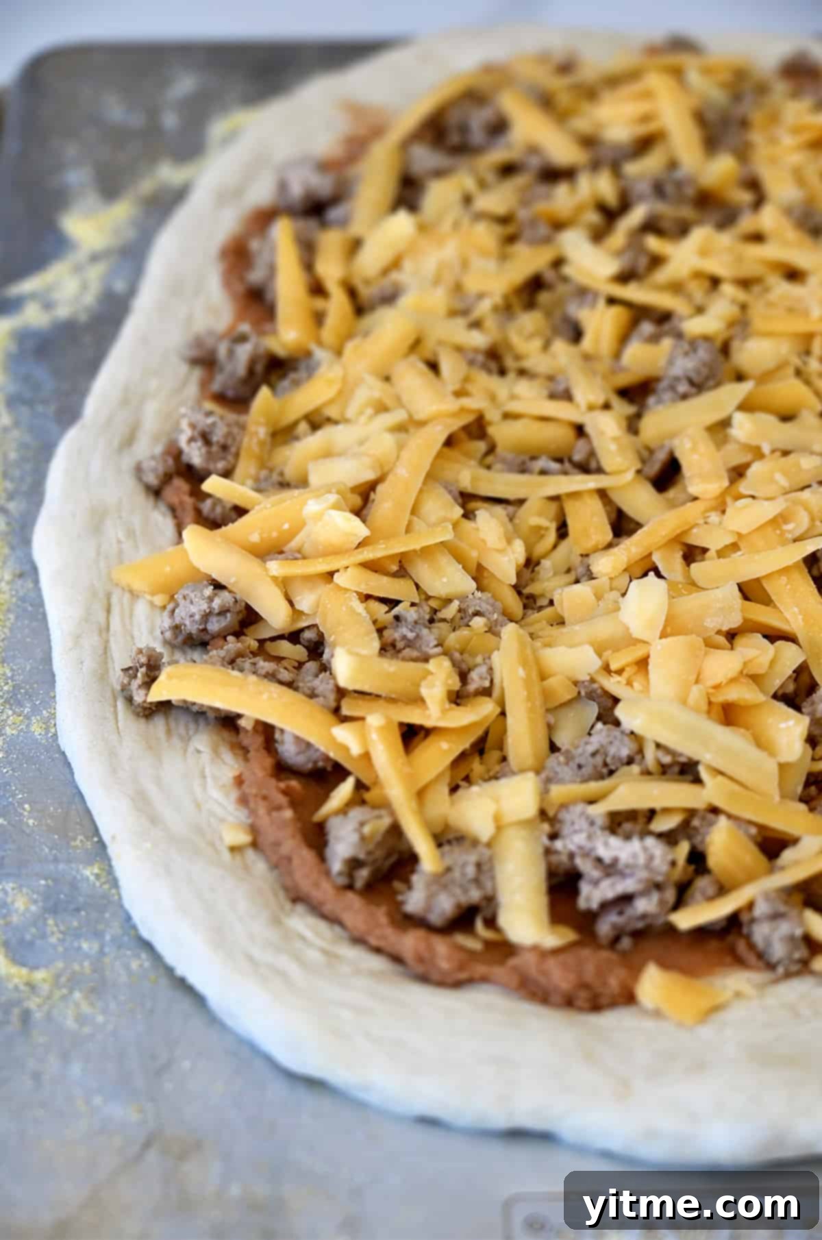 Pizza dough topped with refried beans, cooked taco meat and shredded cheddar cheese.