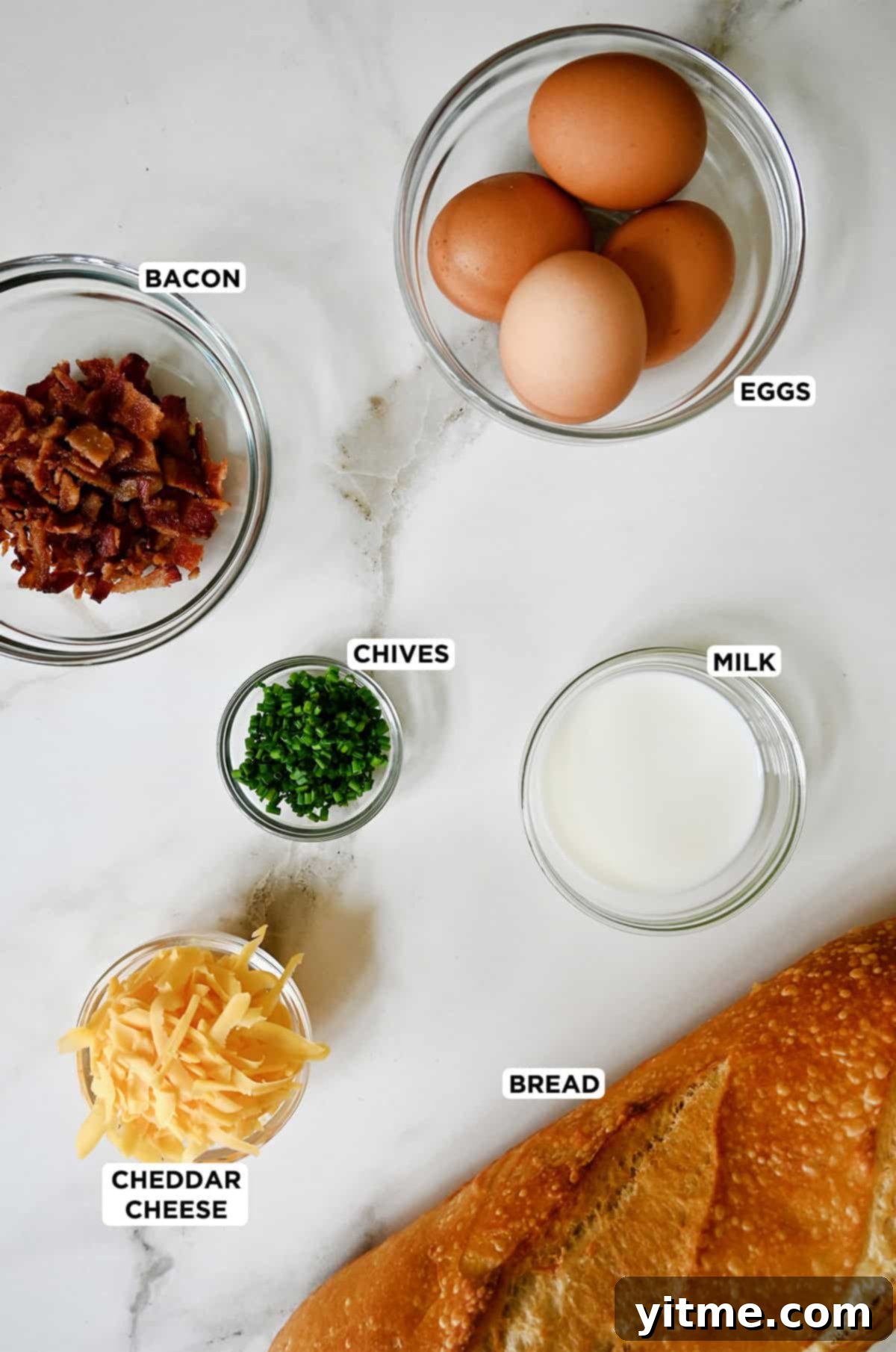 A beautifully arranged spread of sourdough bread, shredded cheddar cheese, chopped fresh chives, cooked crumbled bacon, fresh eggs, and a small jug of milk, all ready for making cheesy baked egg boats.