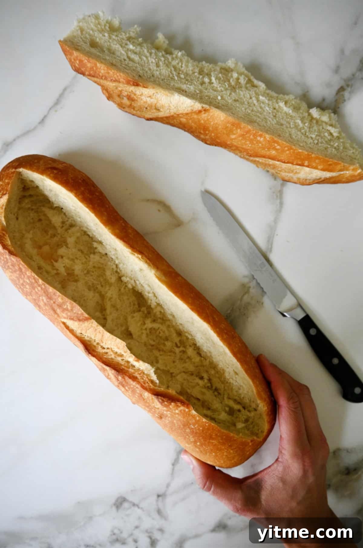A hand gently holds a sourdough loaf with a neatly carved "V" channel in its center, showing the hollowed-out space ready for filling. A sharp knife and the removed bread portion are visible nearby on a wooden cutting board.