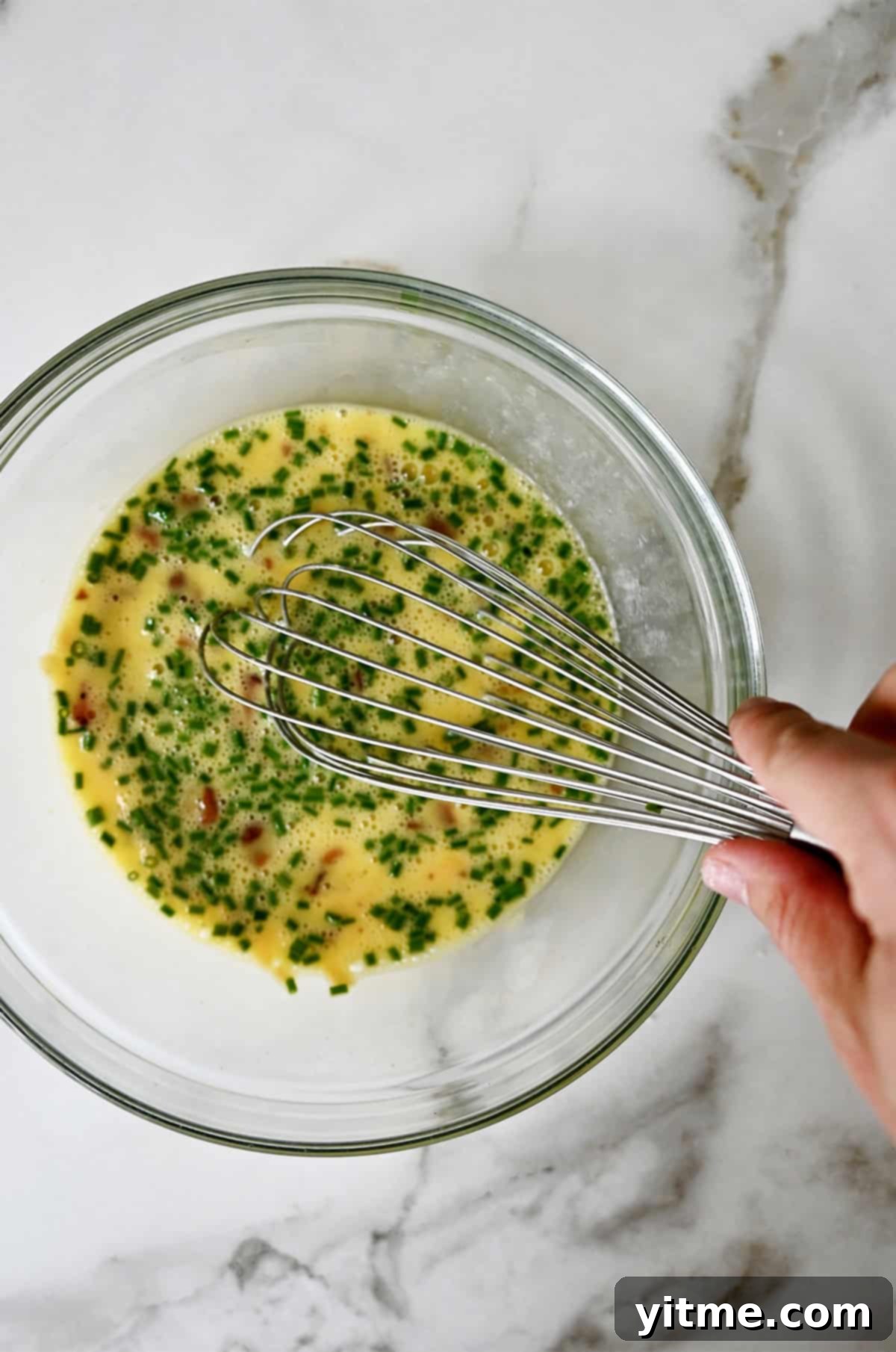 A close-up of a hand whisking eggs, milk, shredded cheese, and crumbled bacon in a bowl, creating the delicious filling for the baked egg boats.