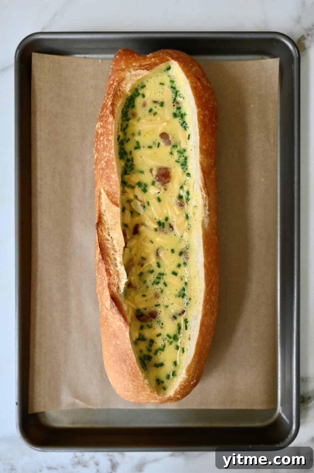 An unbaked sourdough bread boat, generously filled with a creamy scrambled egg, cheese, and bacon mixture, sitting on a parchment paper-lined baking sheet, ready for the oven.