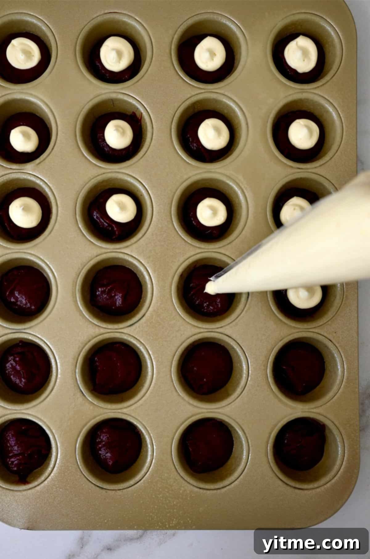 Piping cheesecake filling atop red velvet batter in a mini muffin tin.