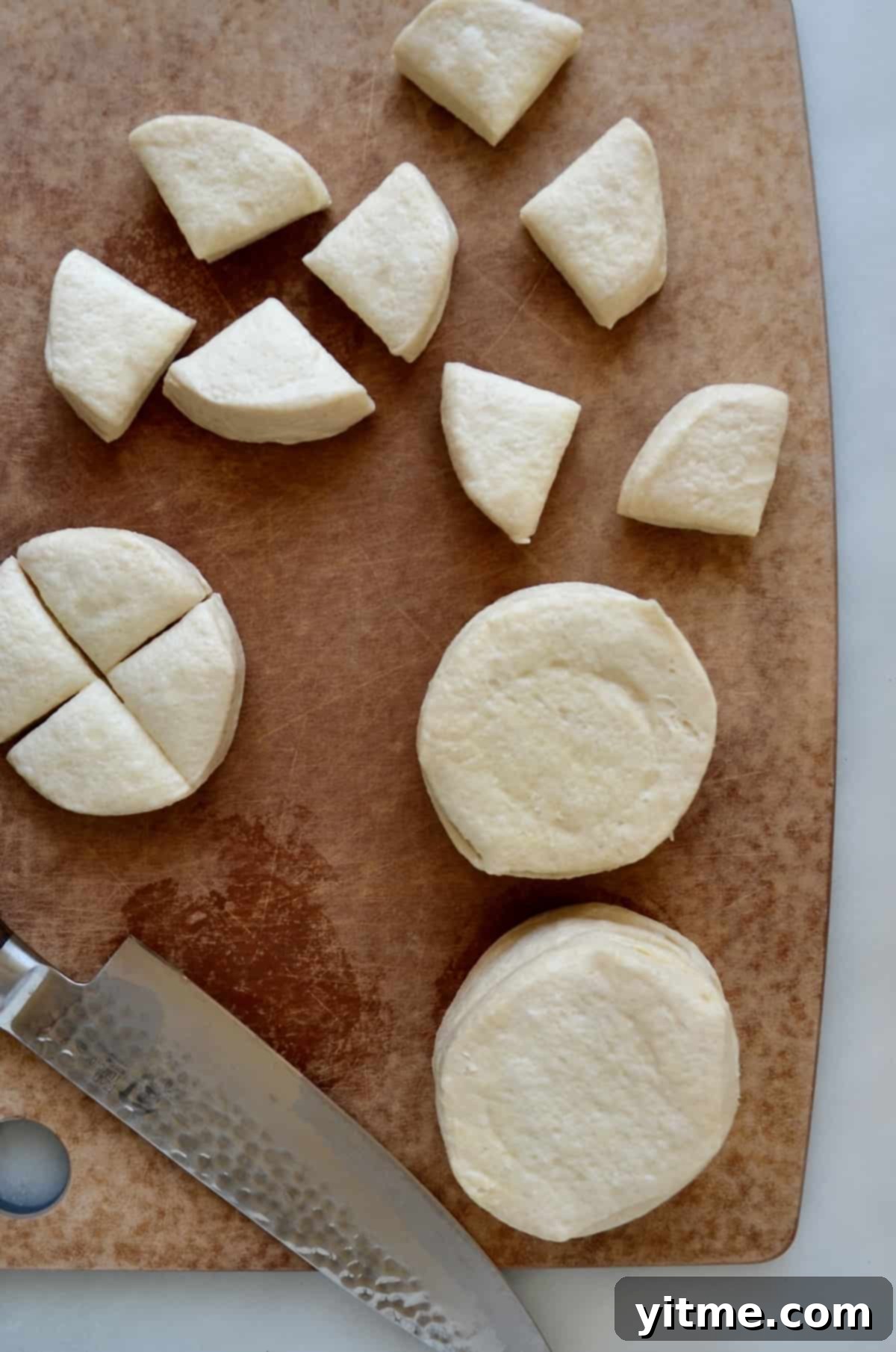 Canned biscuit dough being cut into pieces on a wooden cutting board with a chef's knife.
