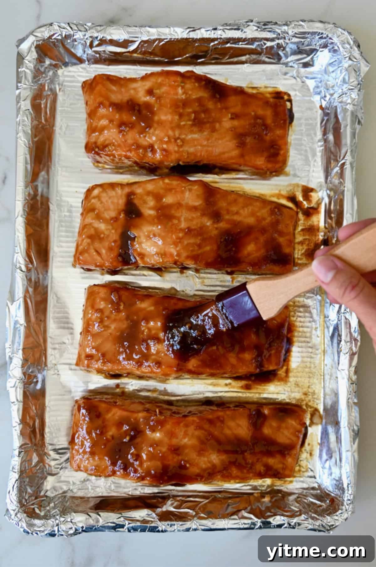 Honey-Garlic Glazed Salmon Delight 8 A hand holding a brush applies a honey garlic glaze to oven-baked salmon filets on a baking sheet lined with foil.