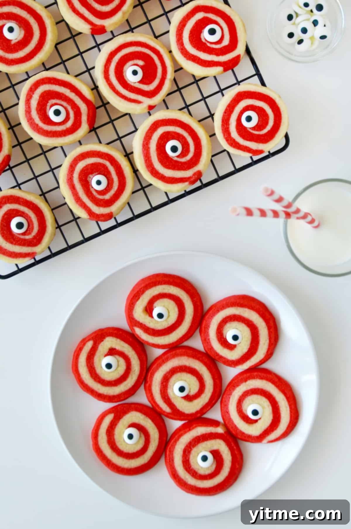 A vibrant plate of red and white Halloween pinwheel cookies, each adorned with a fun monster eye, resting beside a cool glass of milk. More freshly baked cookies are visible on a cooling rack in the background.