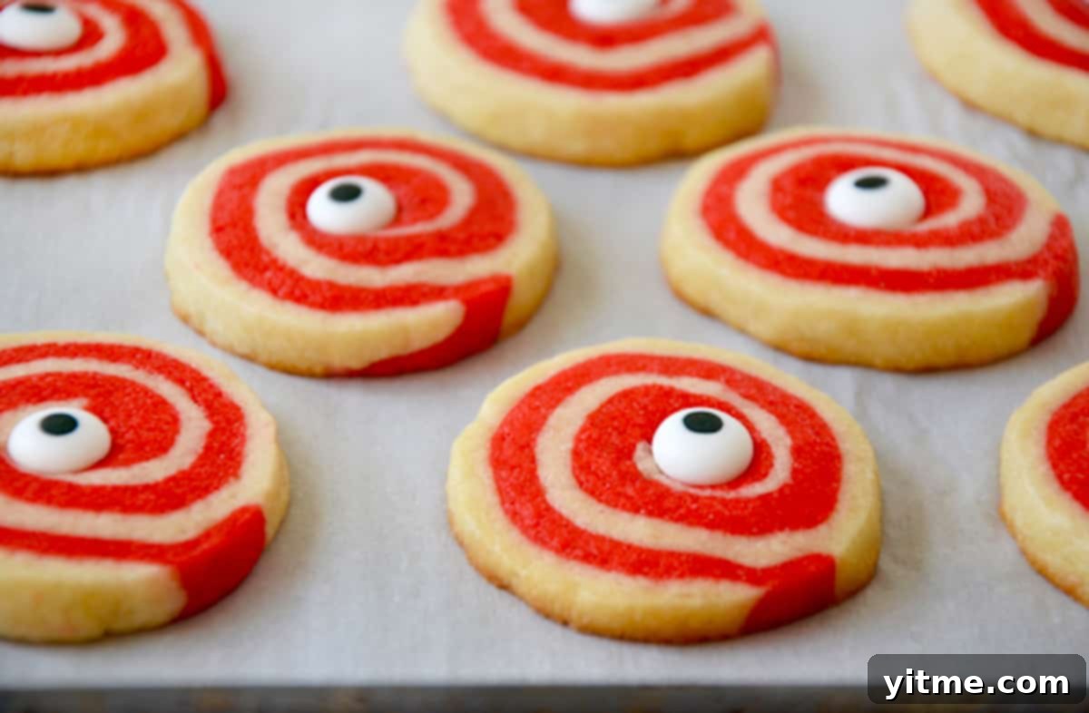 A baking sheet covered with parchment paper holds several freshly baked red and white pinwheel cookies, each adorned with a whimsical candy eyeball in its center, ready to cool and be enjoyed.