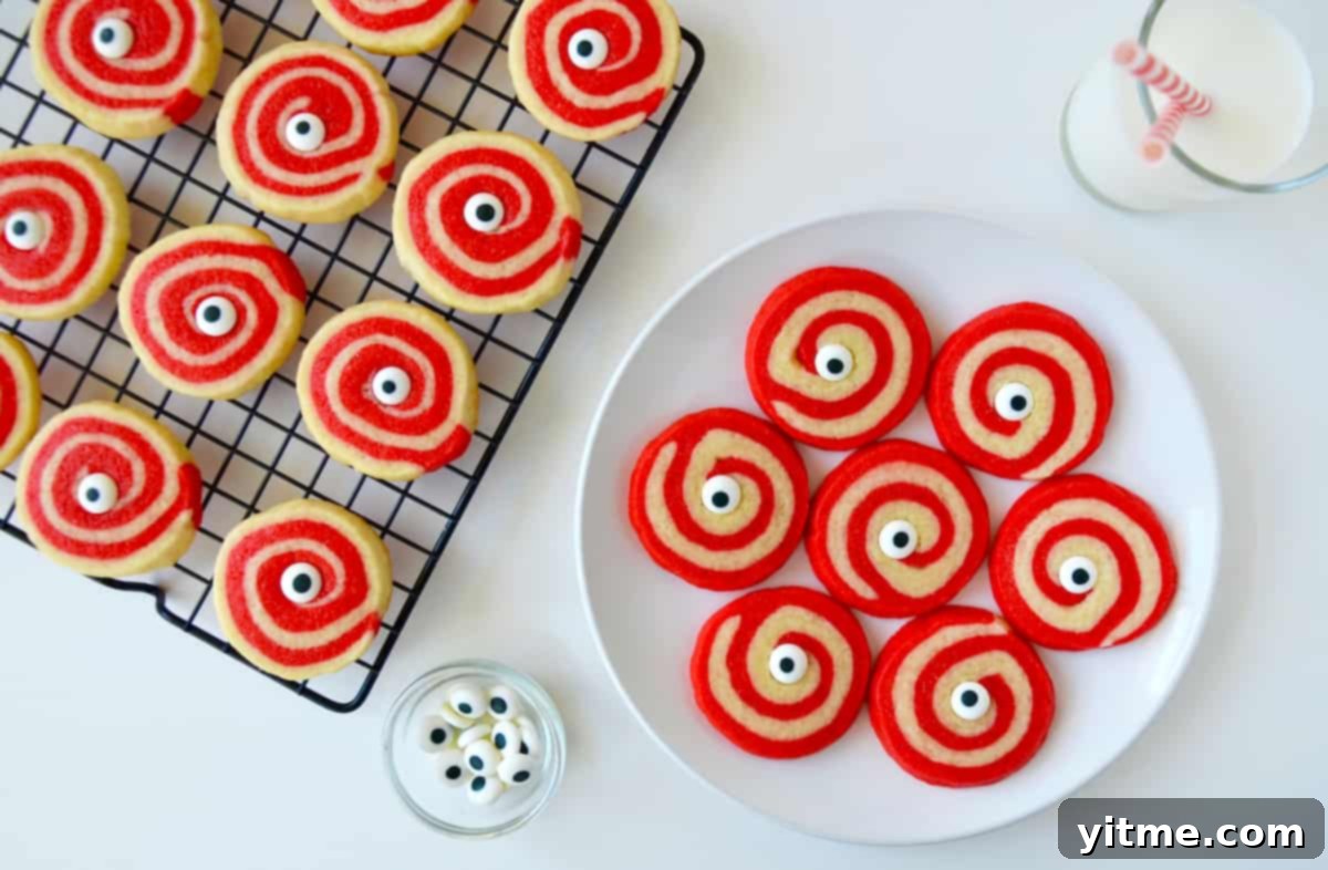 A charming scene of Halloween sugar cookies, each a red and white pinwheel adorned with a candy eyeball, neatly arranged on a plate. A small bowl of extra candy eyeballs, a refreshing glass of milk, and more cookies cooling on a wire rack complete this festive display.