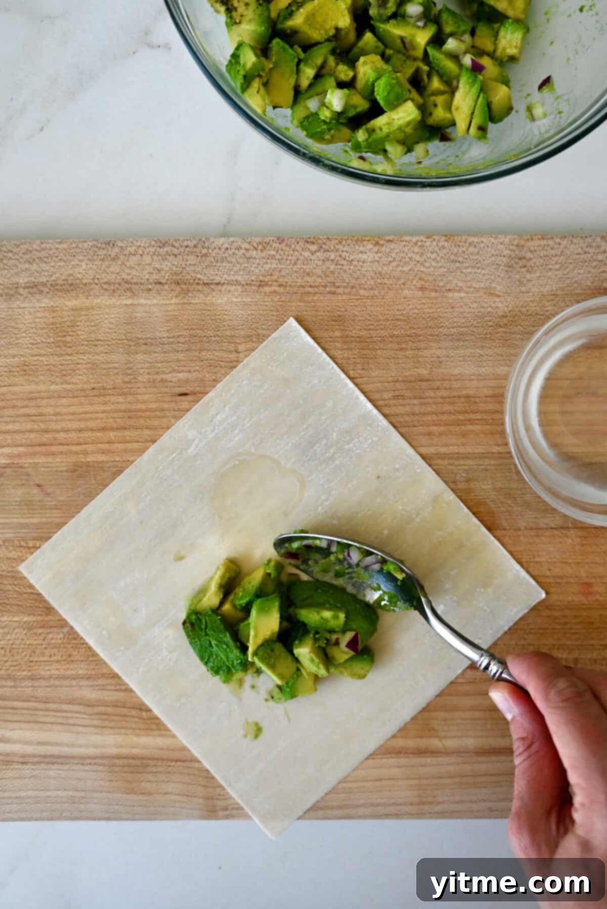 Avocado filling being spooned atop an egg roll wrapper that's on a wood cutting board along with a small glass bowl filled with water.