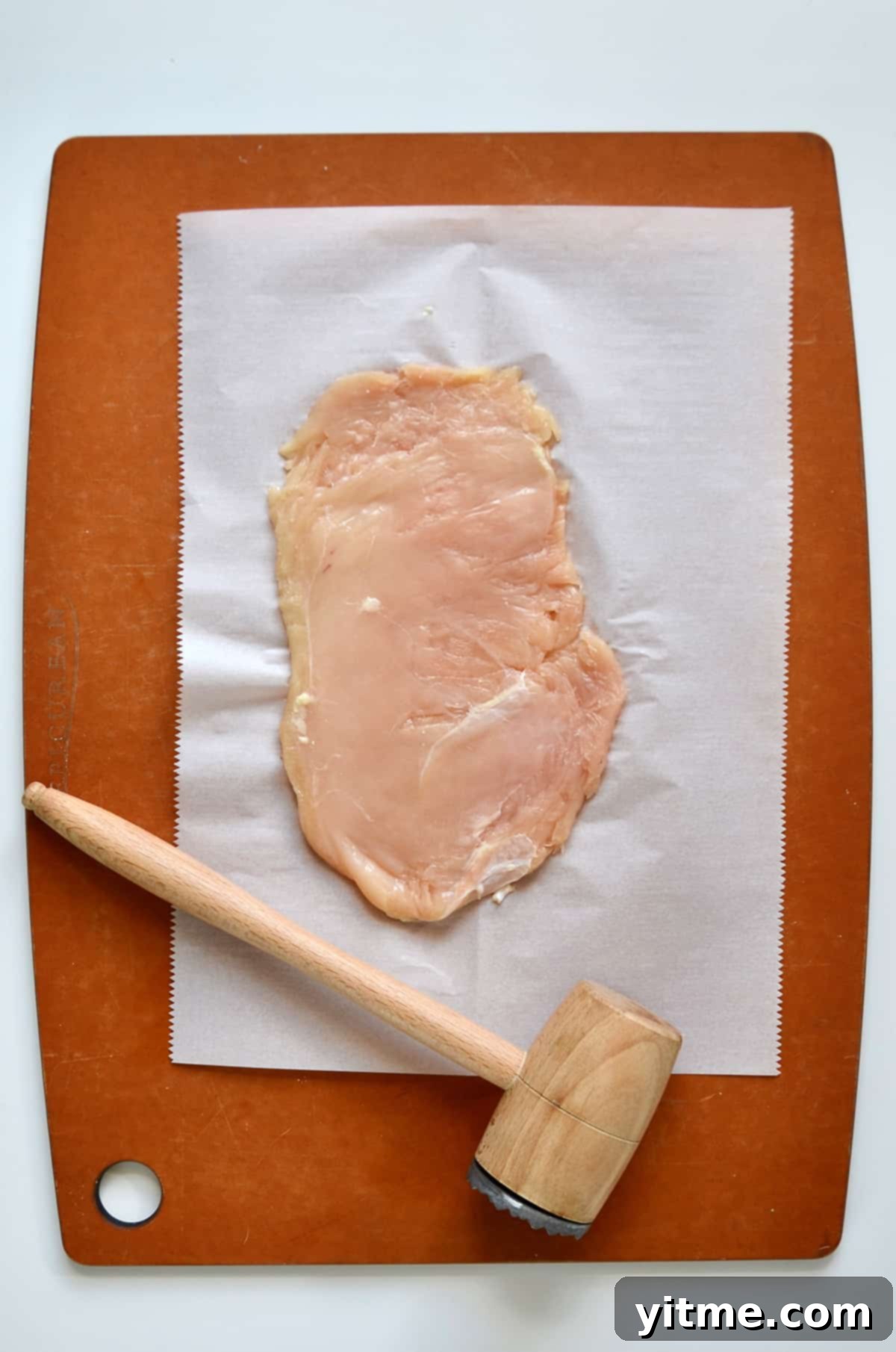 A piece of thinly pounded chicken is on a piece of parchment paper on a cutting board, with a wooden meat mallet nearby.