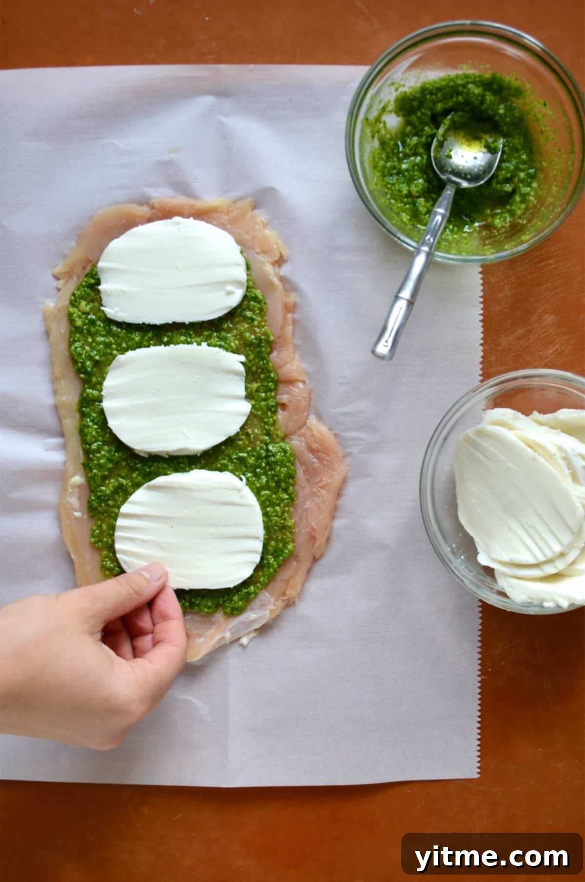 Thinly pounded chicken is topped with pesto and sliced mozzarella. Nearby are bowls of pesto and mozzarella cheese.