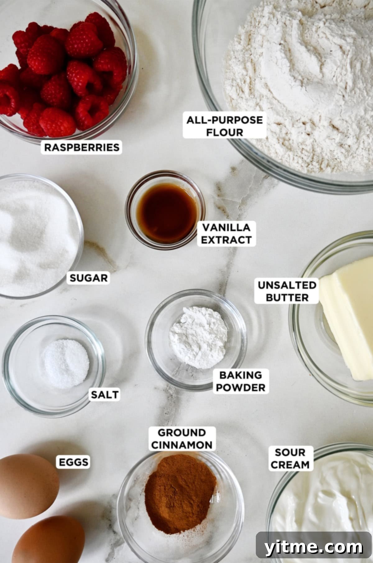 Various sizes of glass bowls containing classic coffee cake ingredients: flour, vanilla extract, butter, baking powder, cinnamon, sugar, salt, eggs, sour cream and raspberries.